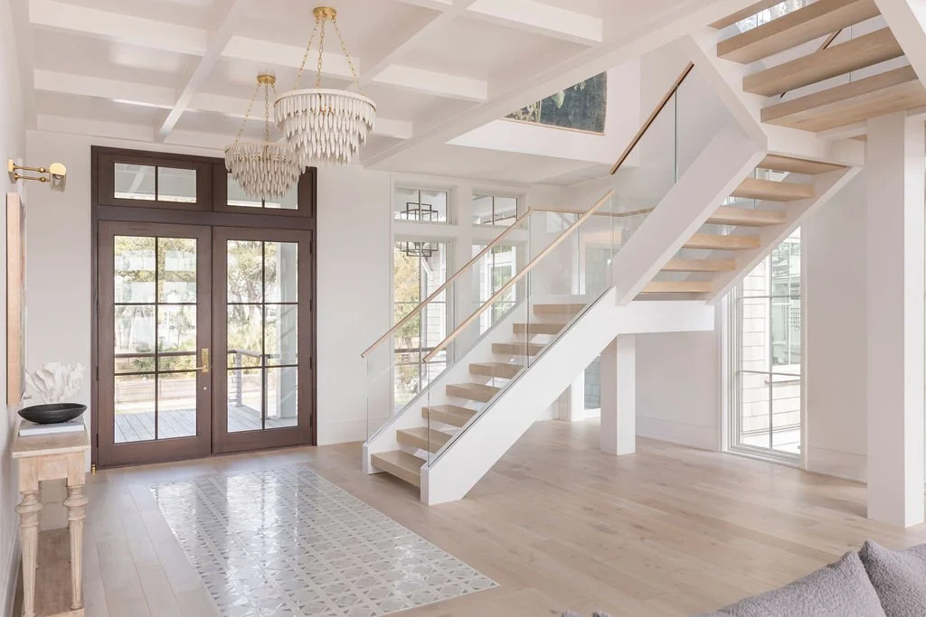 Bright entryway with staircase, glass railings, chandelier, and wooden doors leading outside.