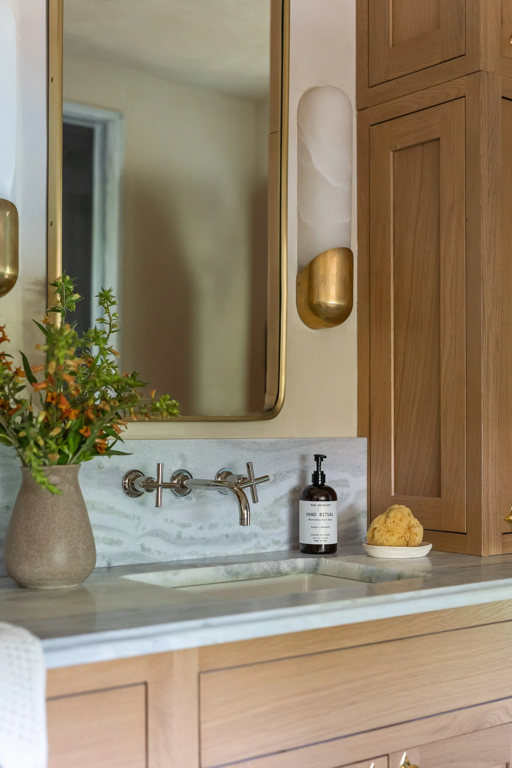 A kitchen countertop with a beige vase holding green and orange flowers, a soap dispenser labeled 'HAND RITUAL,' a sponge on a white dish, with wooden cabinets, a mirror, a window, and decorative gold wall sconces in the background.