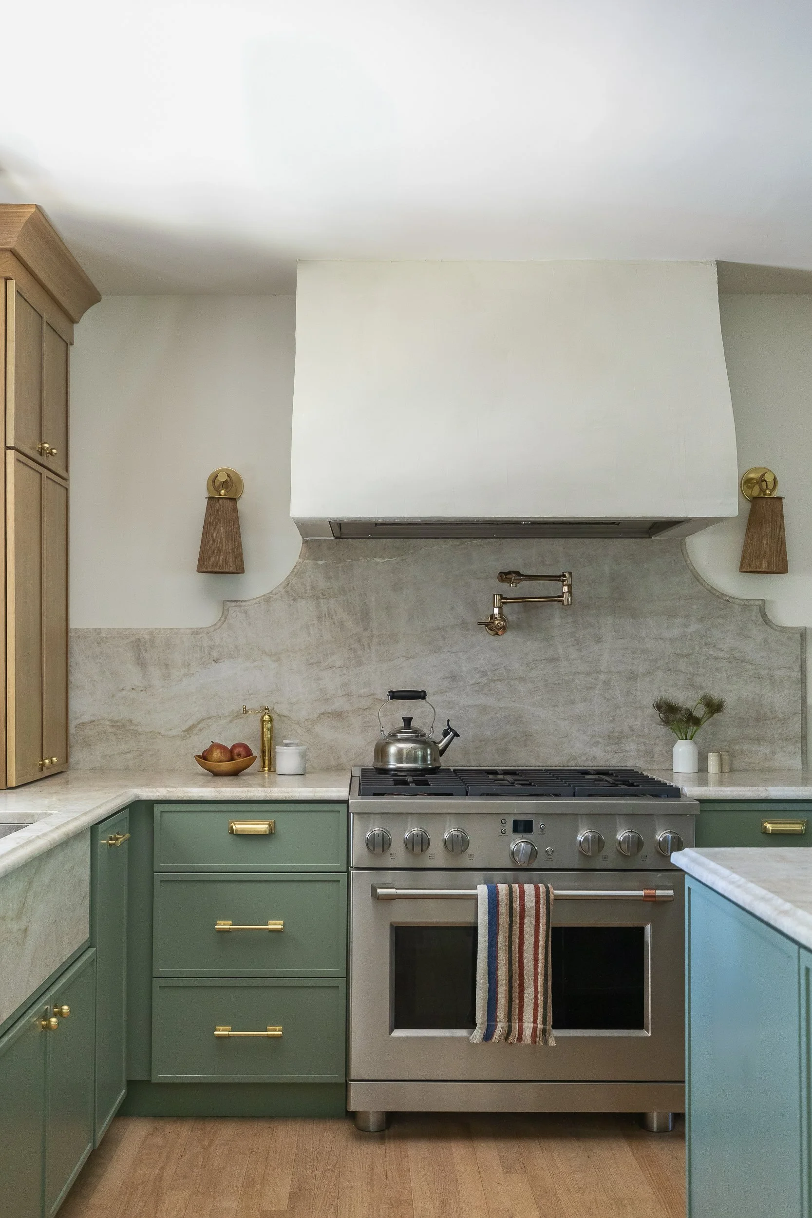 Kitchen with green cabinets, a stainless steel stove, a teapot on the stove, and a white range hood. Decor includes a bowl of apples, a white container, and a small vase with greenery. Wall-mounted lamps with brown shades are next to the range hood.