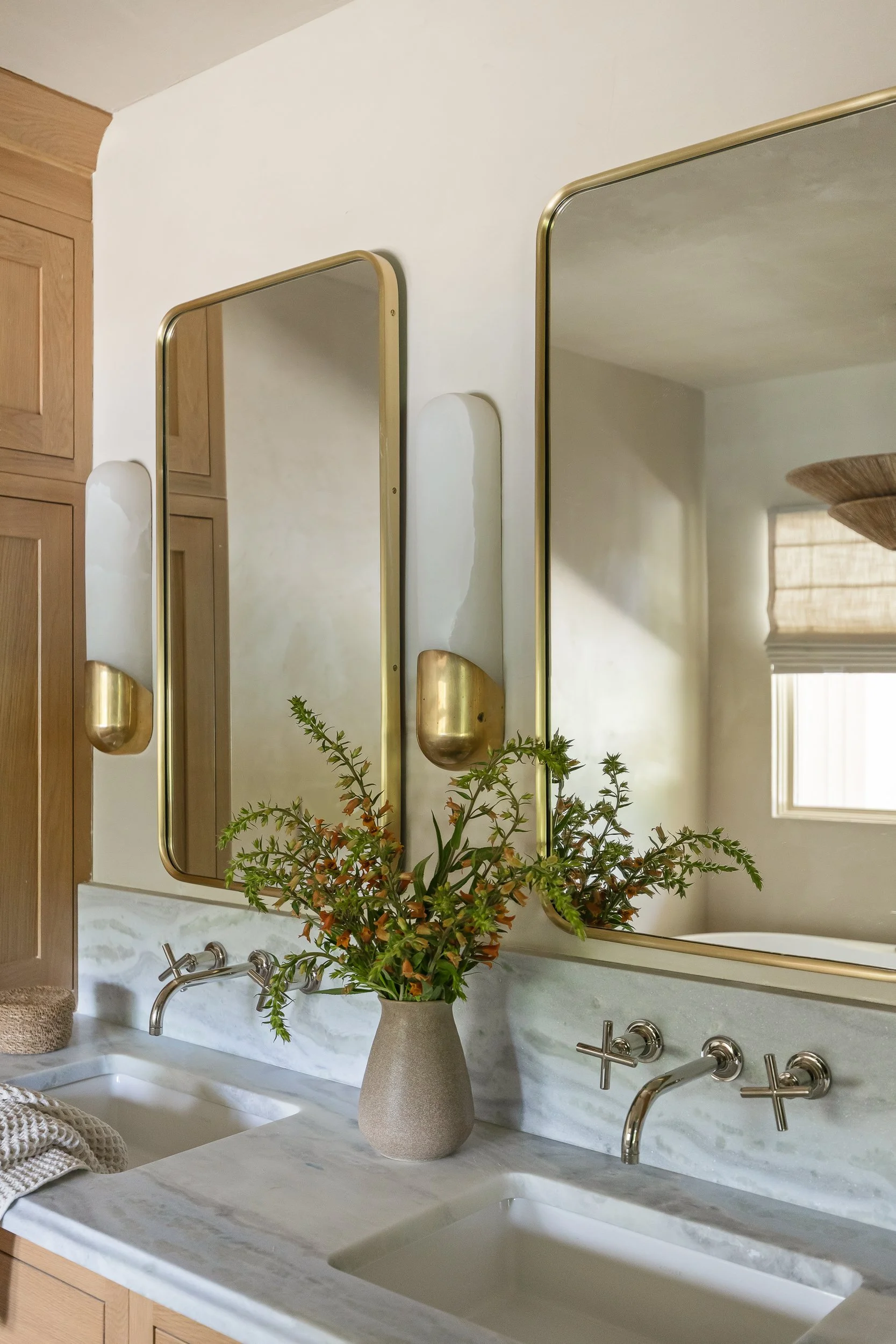 Bathroom sink with a marble countertop, a large mirror, a beige flowerpot with green and orange foliage, and a wooden cabinet with closed doors.