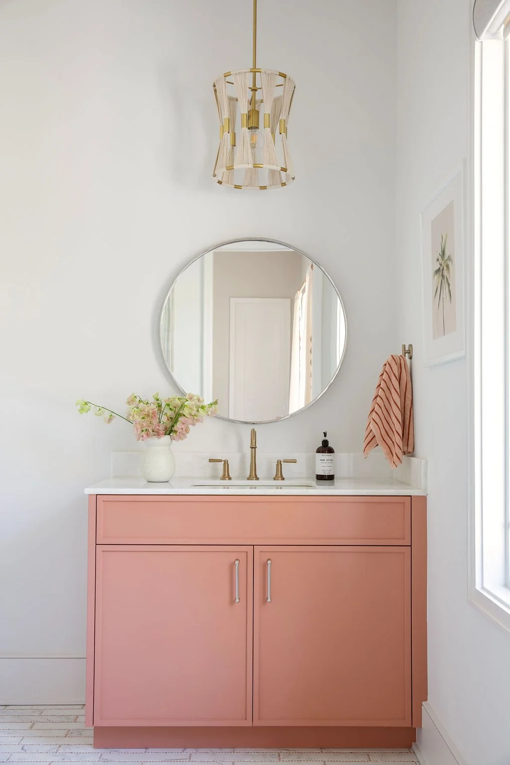 A bathroom vanity with a pink cabinet, gold faucet, and a large round mirror. A vase with pink flowers is on the left, a soap dispenser is on the right, and a pink towel hangs on the wall. A white wall, a framed picture, and a window are in the backg