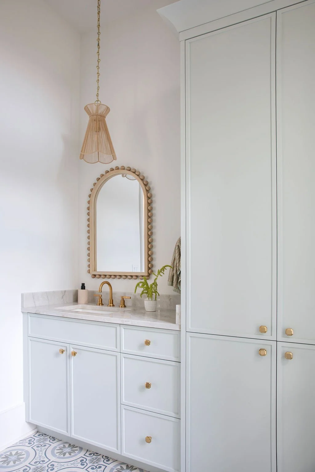 Bathroom with white vanity, marble countertop, gold fixtures, an oval mirror with a wooden bead frame, a hanging light fixture, a potted fern, and a built-in closet with white doors.