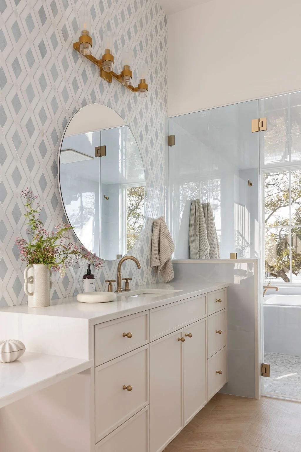 Bathroom with white vanity, oval mirror, gold faucet, patterned wallpaper, towel hooks with beige towels, glass shower enclosure, bathtub, and window designed by JLV Creative, built by Scenic Custom Homes and styled by Styled by Ellen Casey