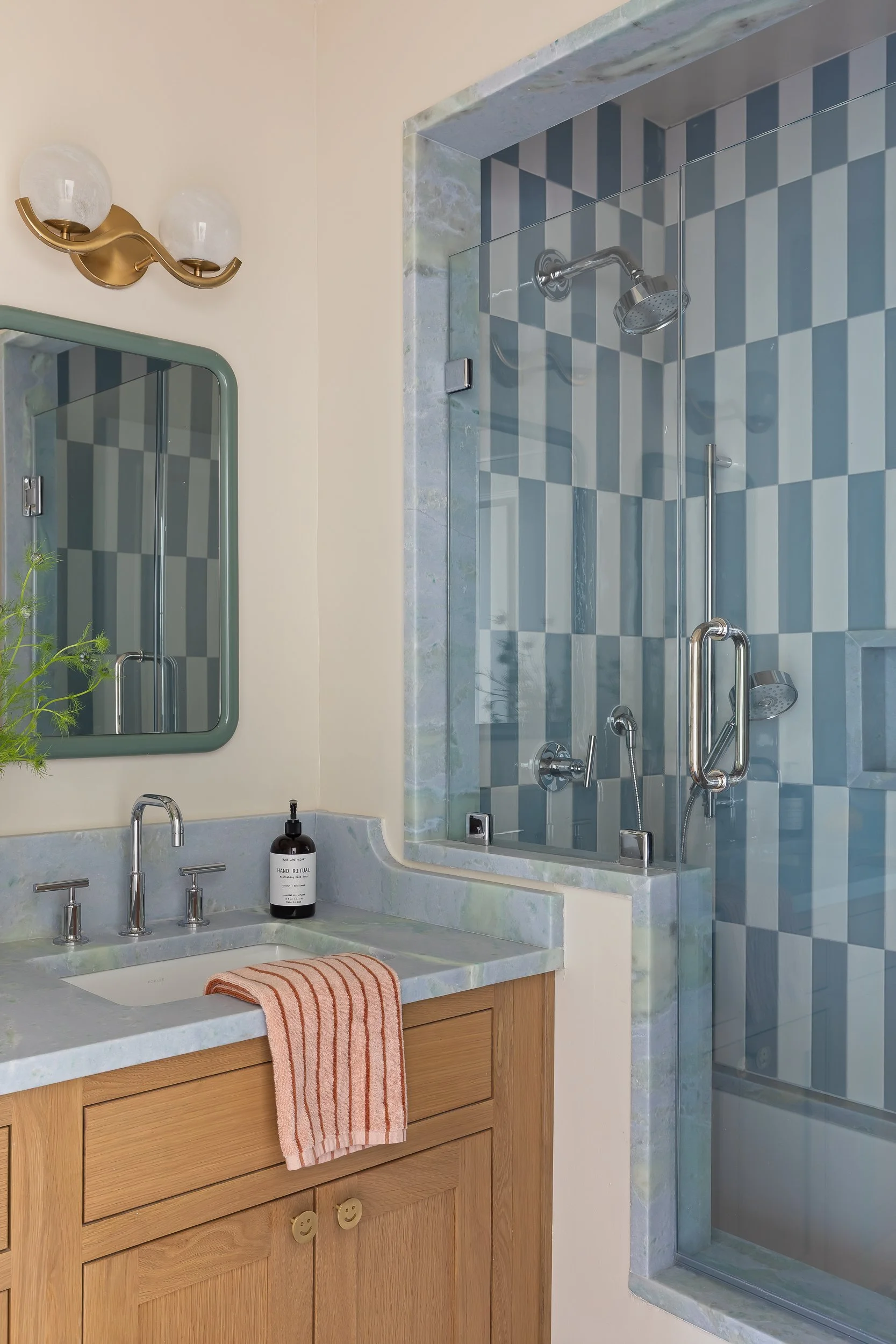 Bathroom vanity with wooden cabinet, marble countertop, sink, faucet, hand soap dispenser, and pink striped towel; mirror, wall-mounted light fixture, and glass-enclosed shower with blue and gray checkered tiles in the background.