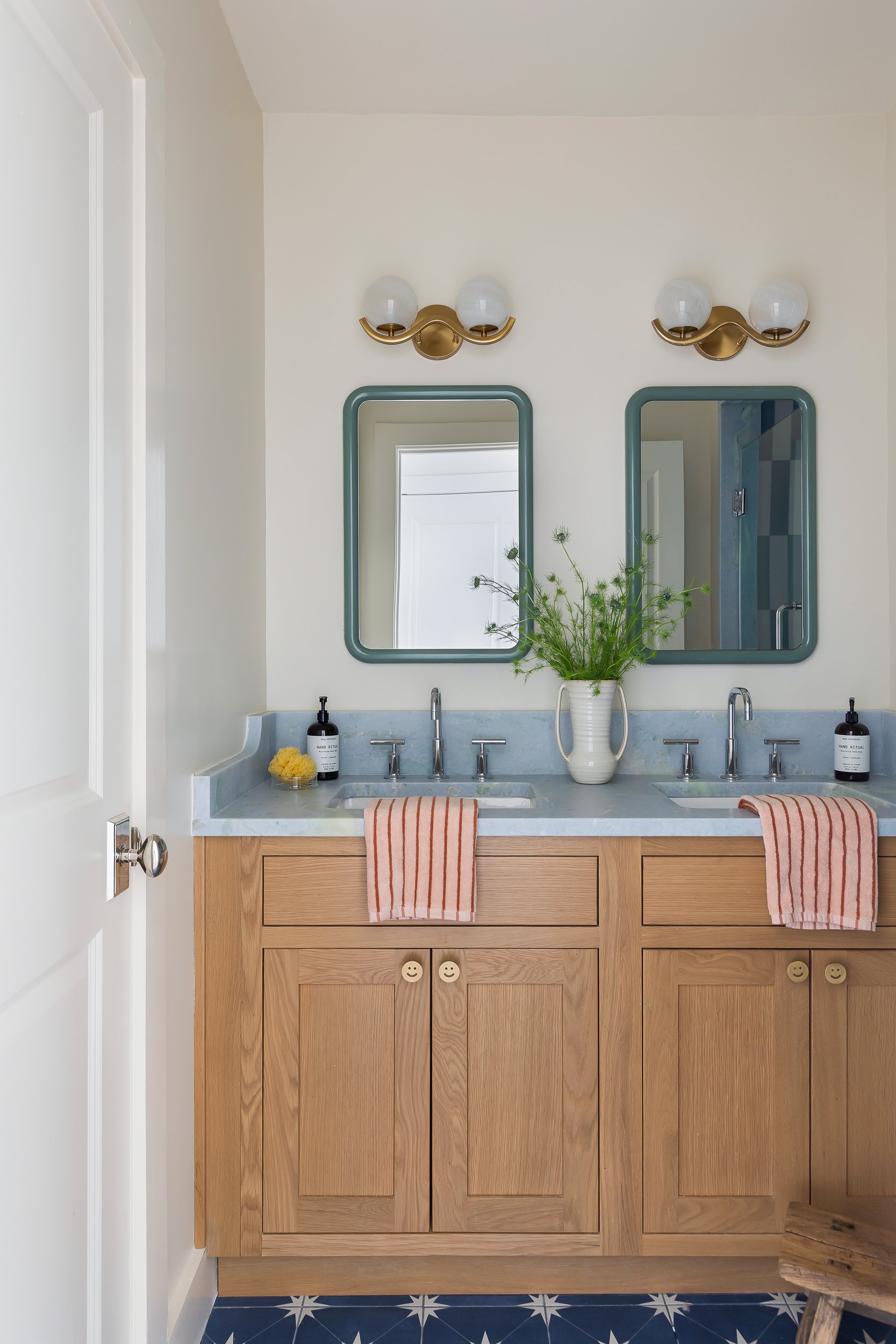 A double bathroom vanity with two rectangular mirrors, a potted plant in the center, and two striped towels hanging on each side. White walls, blue and white star-patterned tile floor, and brass wall-mounted light fixtures above the mirrors.