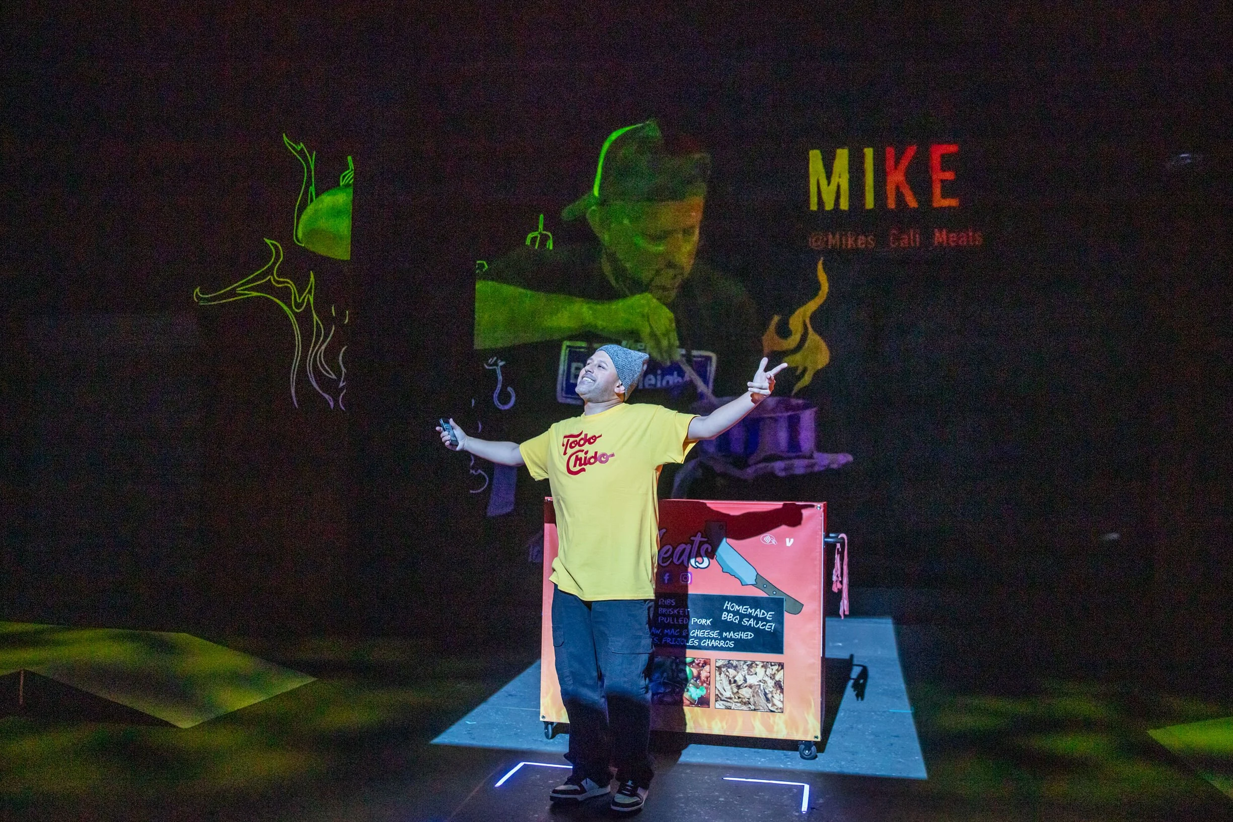 A person in a yellow "Todo Chido" shirt and beanie poses with outstretched arms in front of a colorful projected background. A cart displaying "Mike's Cali Meats" and BBQ menu items is visible. The projection includes abstract designs and a cooking s