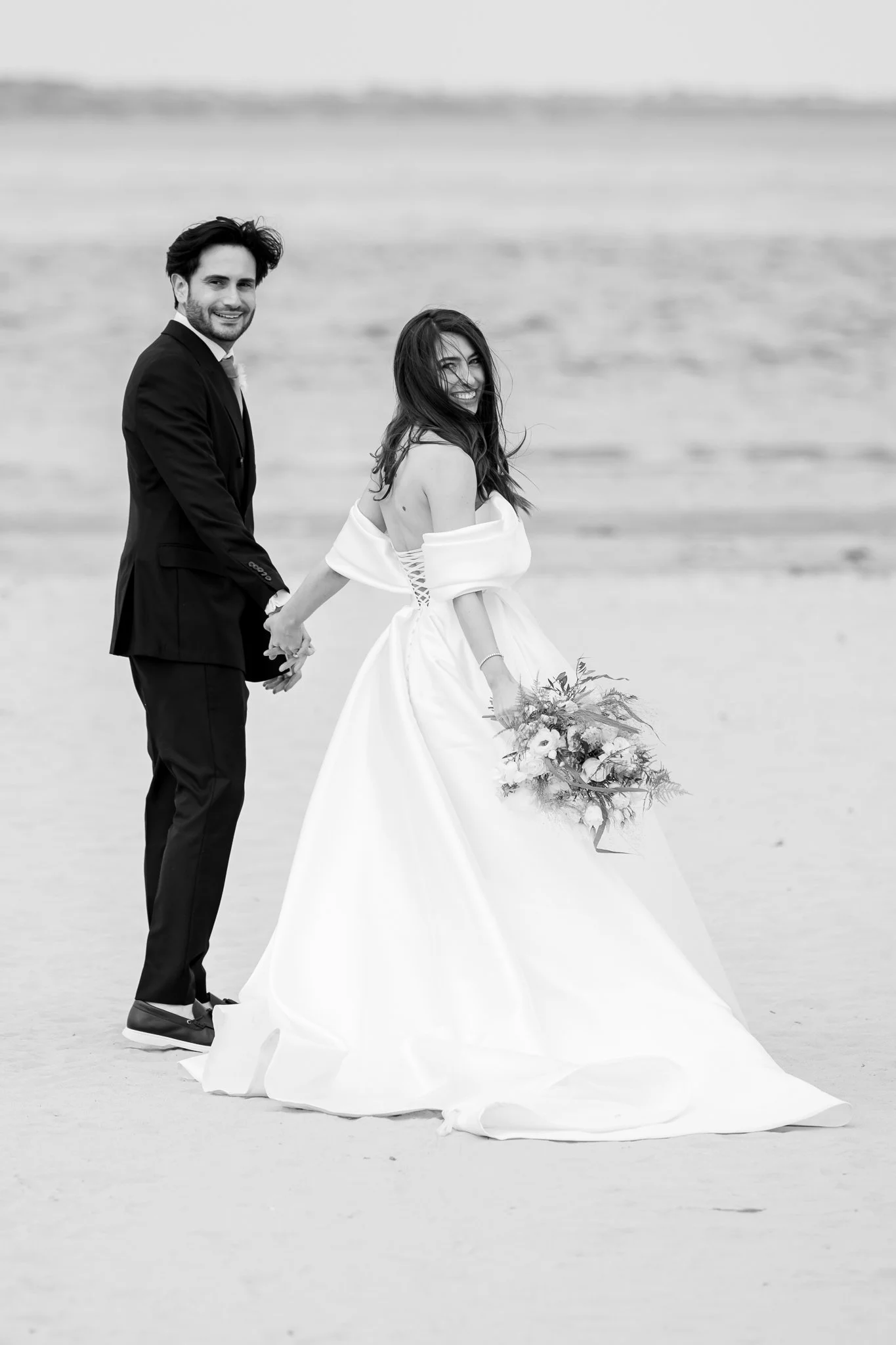 The bride and groom stood on a beach while looking back at the camera man, with the sea in the distance behind them