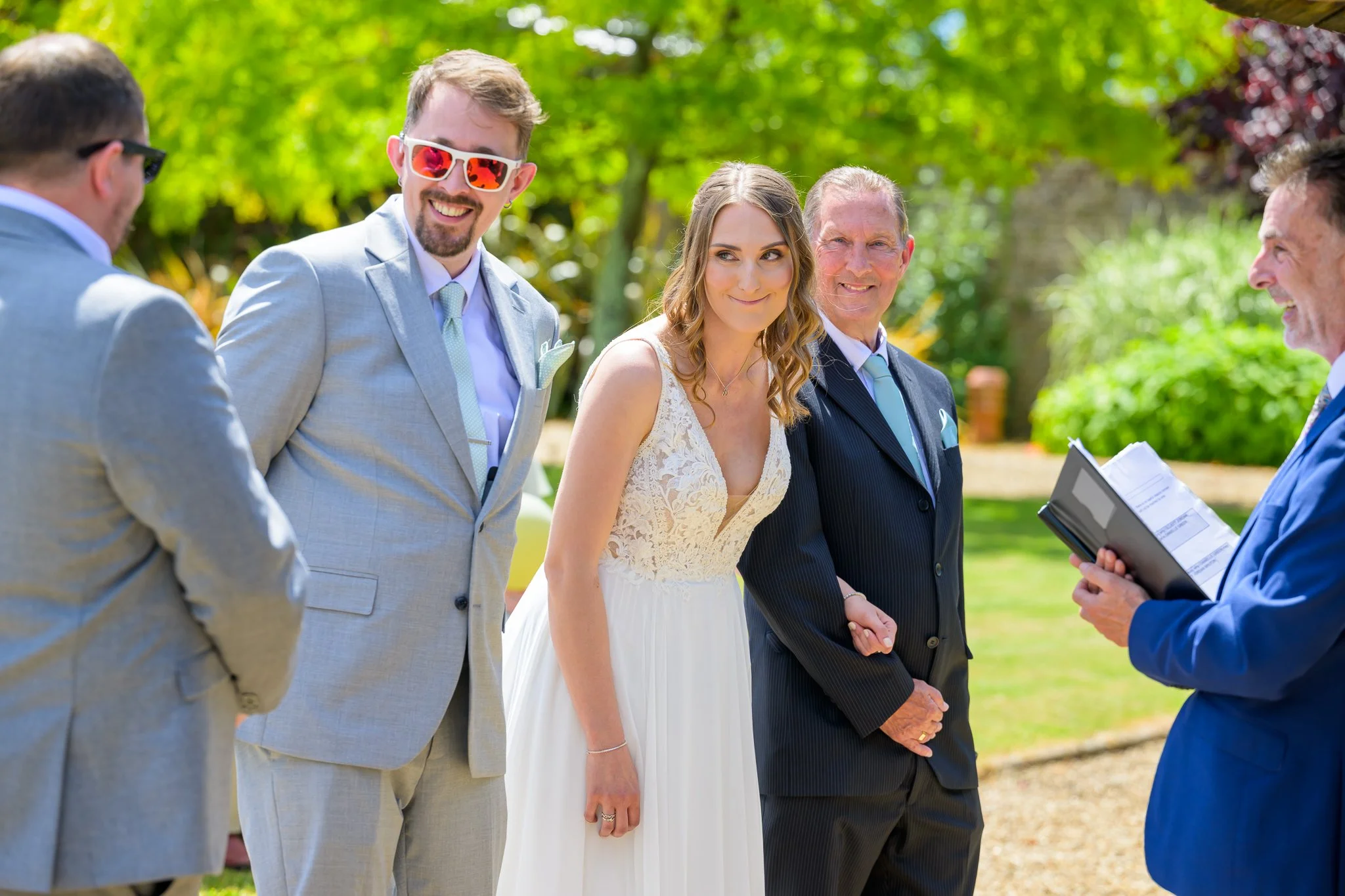 The bride and groom looking at the grooms bestman, while in the middle of saying their wedding vows, with a greenery background