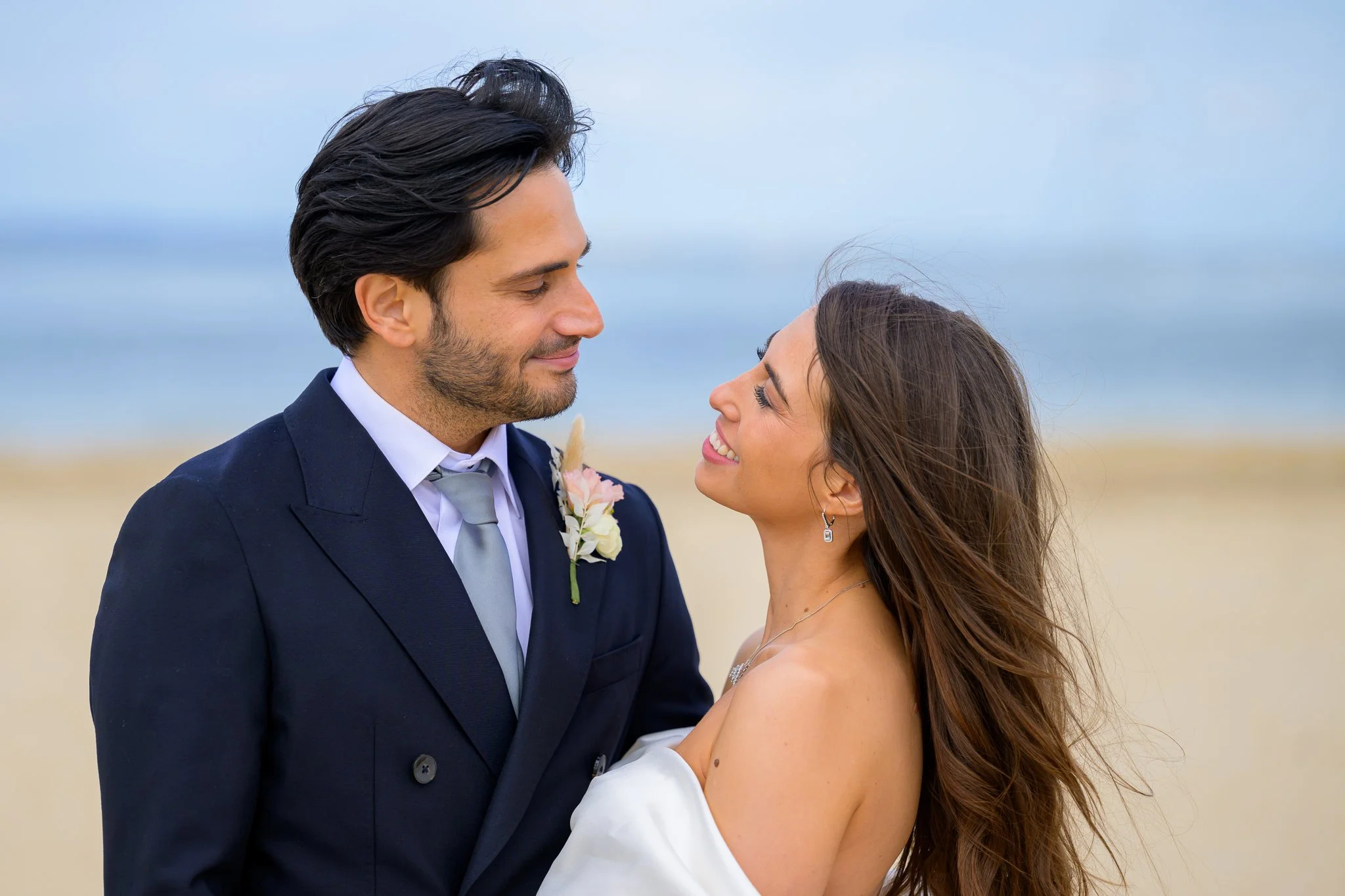 Bride and groom looking at eachother in the eye while stood on a beach, with the ocean and beach behind them