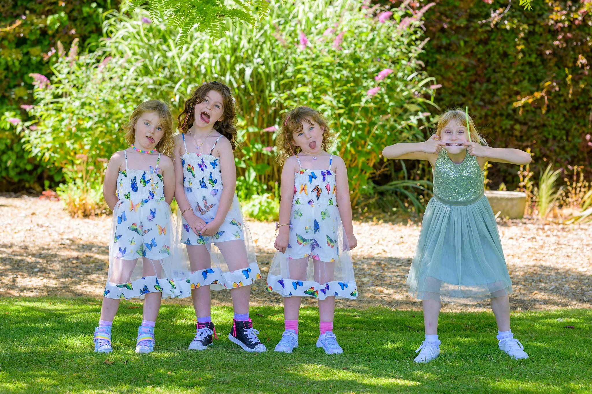 4 children stood on grass posing for the camera, with the 3 children on the left wearing white dresses with colourful spots, and the child on the right wearing a pink dress and making a funny face