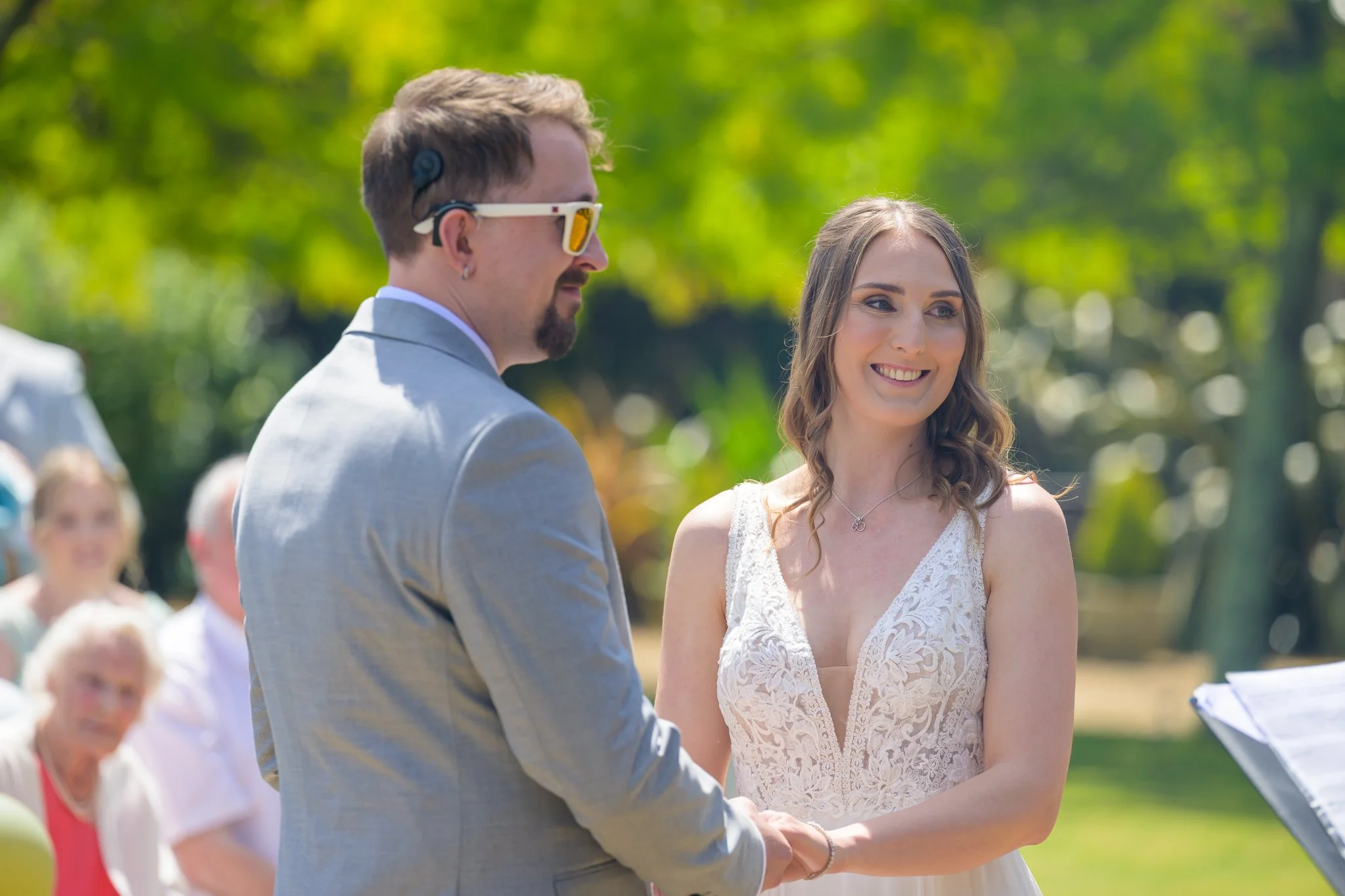 Bride and groom holding hands, looking at the marriage officiant, with trees, bushes, and wedding guests in the background