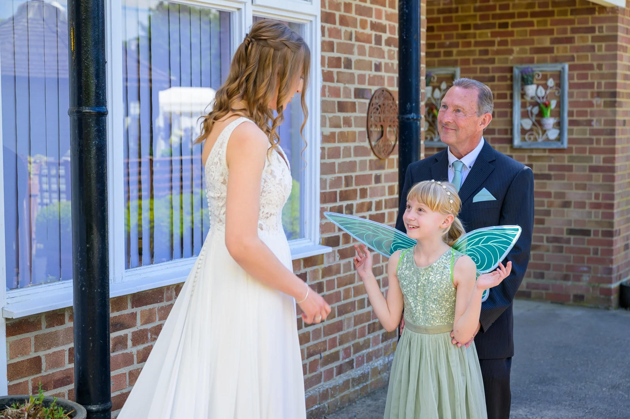 Bride looking at a child wearing a fairy outfit and her father, while stood outside the wedding venue