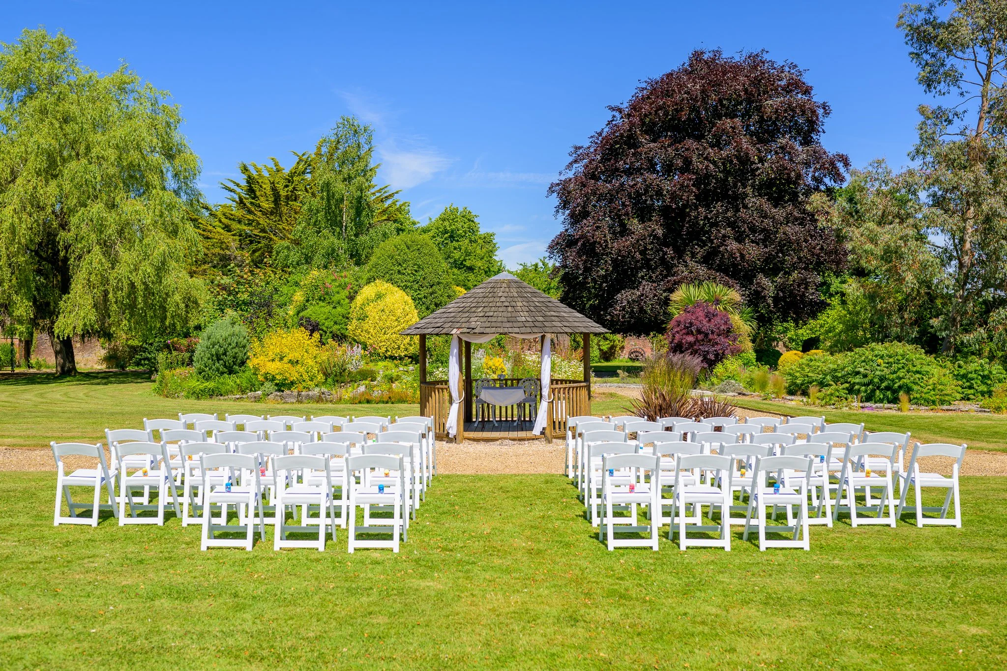 An empty outdoor wedding venue with white seats, and a hut at the end of the aisle, with trees and bushes in the background