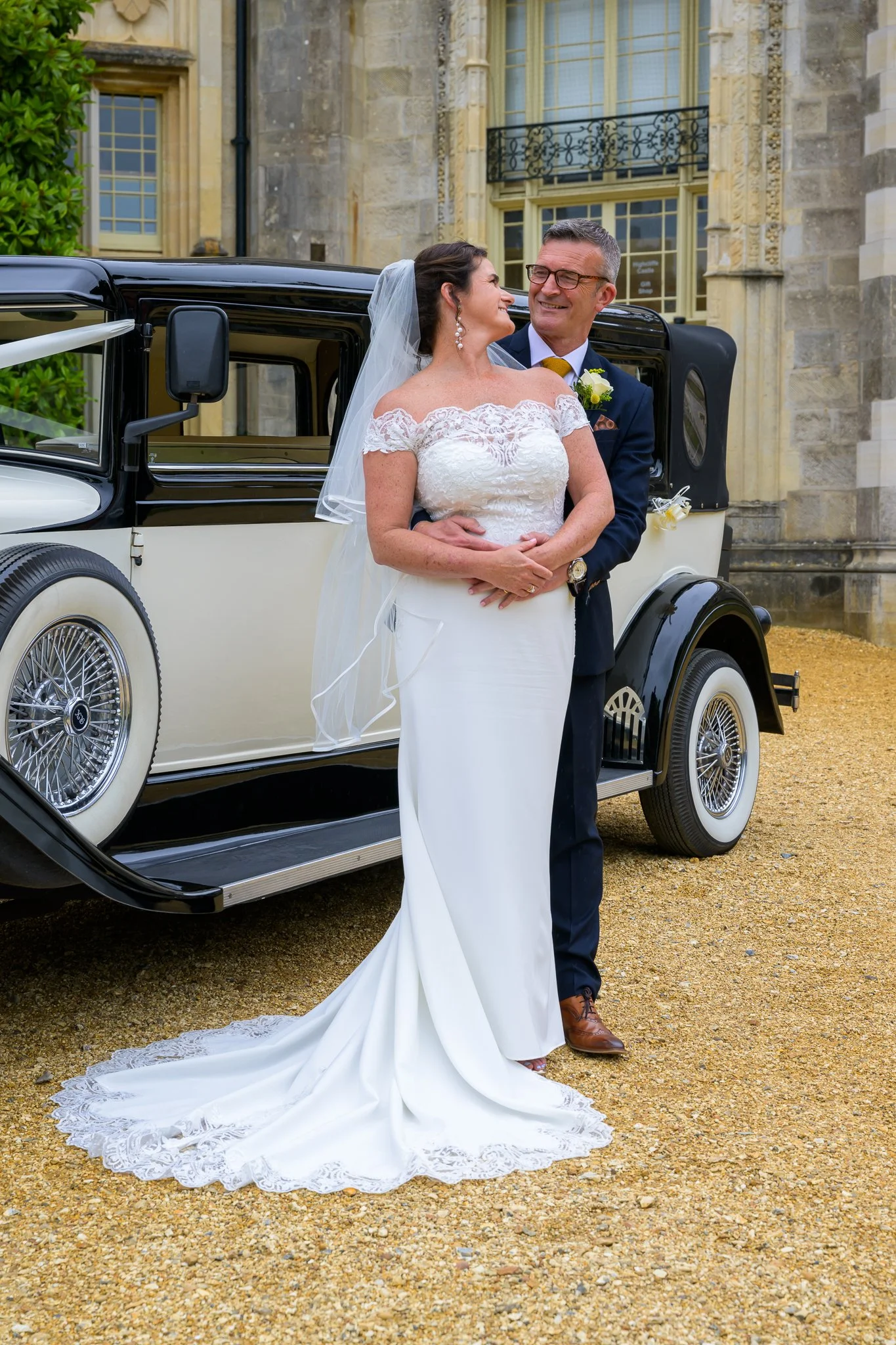 Groom hugging the bride from behind while stood next to their wedding car, outside their wedding venue