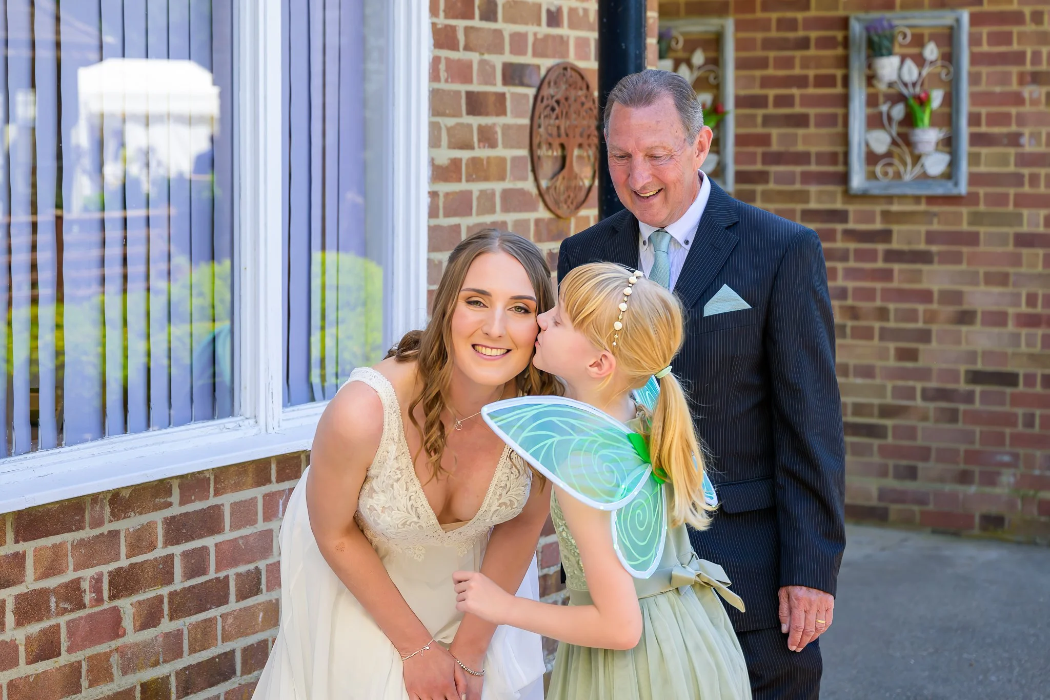 The bride getting kissed on the cheek by a child wearing a fairy outfit, with the brides father and wedding venue behind her