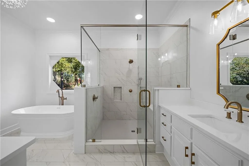 Modern bathroom with white marble walls and floors, glass shower enclosure with gold hardware, white vanity with gold fixtures, a freestanding bathtub, and a window showing green trees outside.