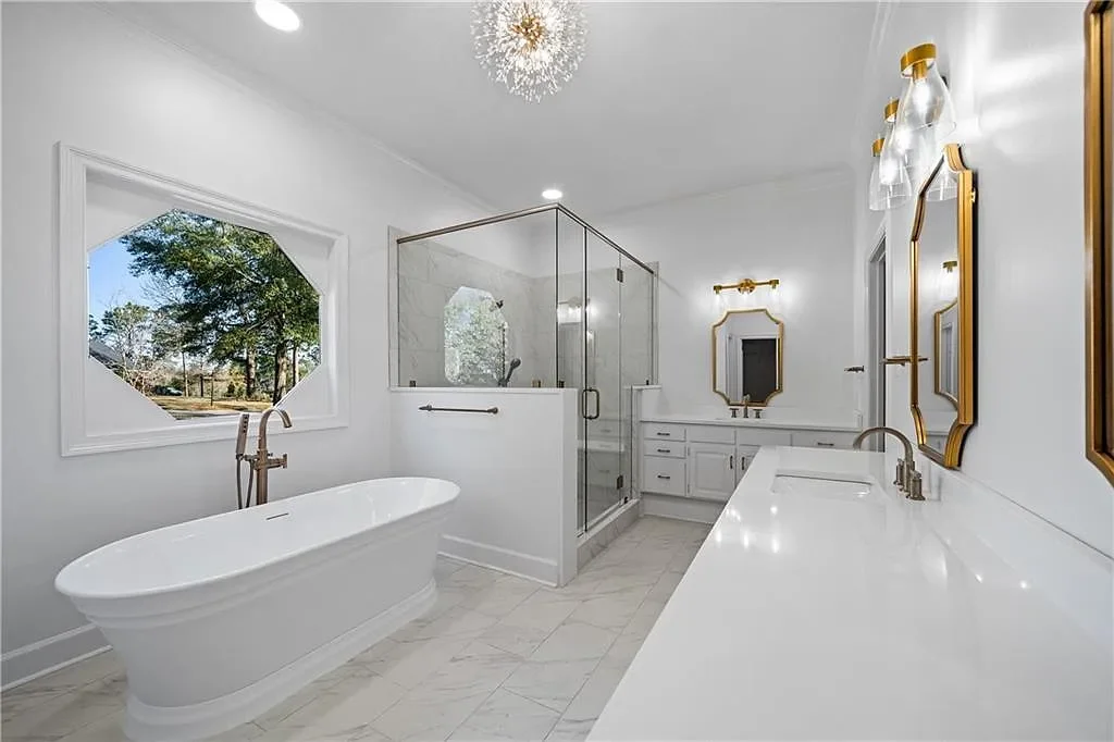 Bright white bathroom with a standalone bathtub, glass-enclosed shower, double vanity with gold fixtures, and a window showing outdoor trees.