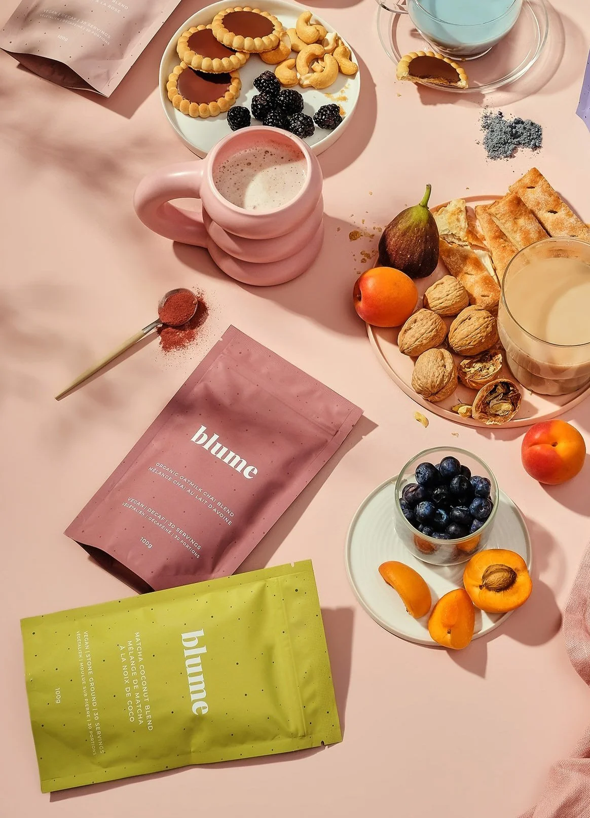 A pink table set with various snacks and beverages. There are plates of cookies, blackberries, walnuts, and apricots. A pink mug holds a frothy drink, and a glass cup with a cookie inside holds a beverage. There are two packets labeled 'blume' with different colors, and a small bowl of blueberries. A spoon with a reddish powder and a pink napkin are also on the table.