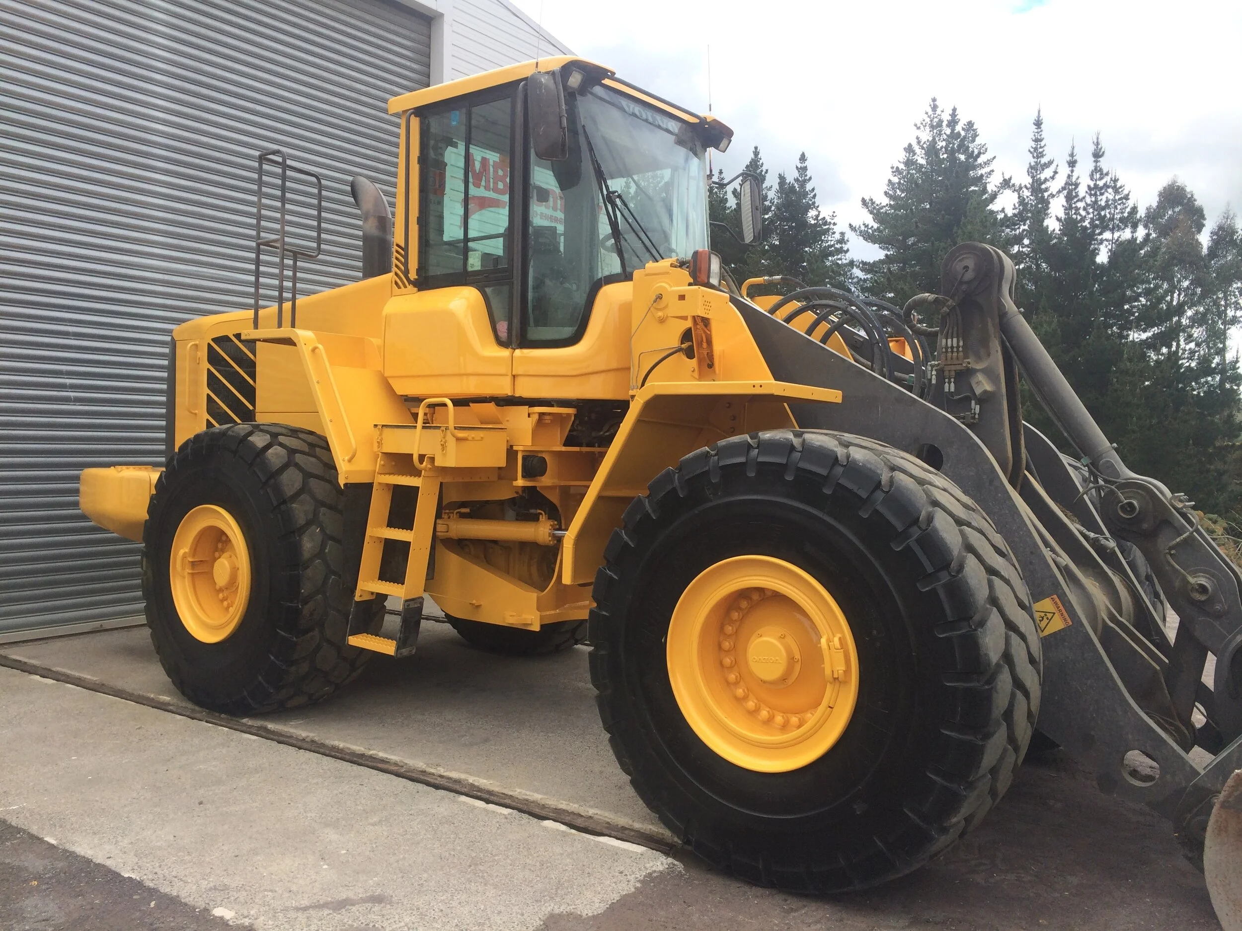 Large yellow construction loader with large wheels, parked on concrete near a metal roller door, surrounded by trees.
