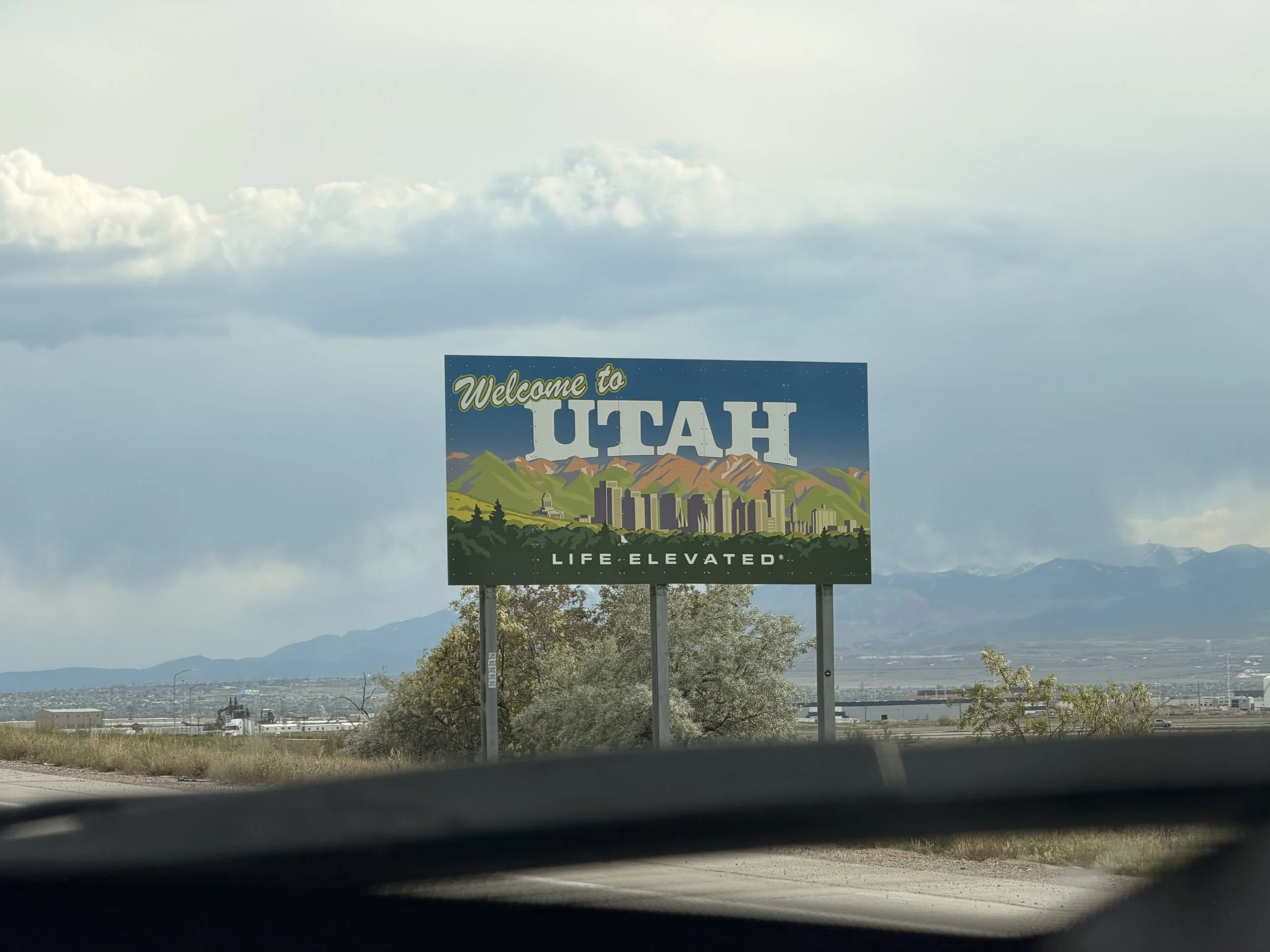 A colorful sign reading "Welcome to Utah" with illustrated mountains and cityscape, located next to the road through a semi-arid landscape with mountains in the background under a partly cloudy sky.