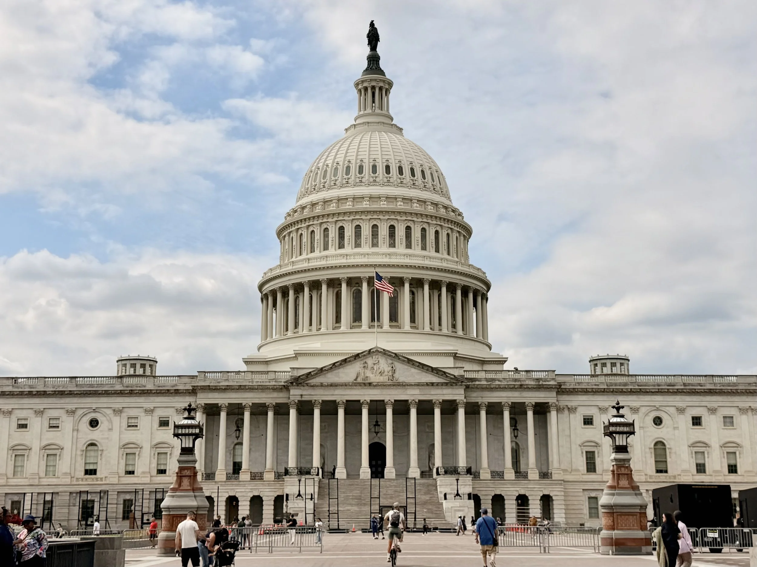 The Capitol building in Washington D.C. with a flag in front, visitors walking and taking photos, and a cloudy sky overhead.