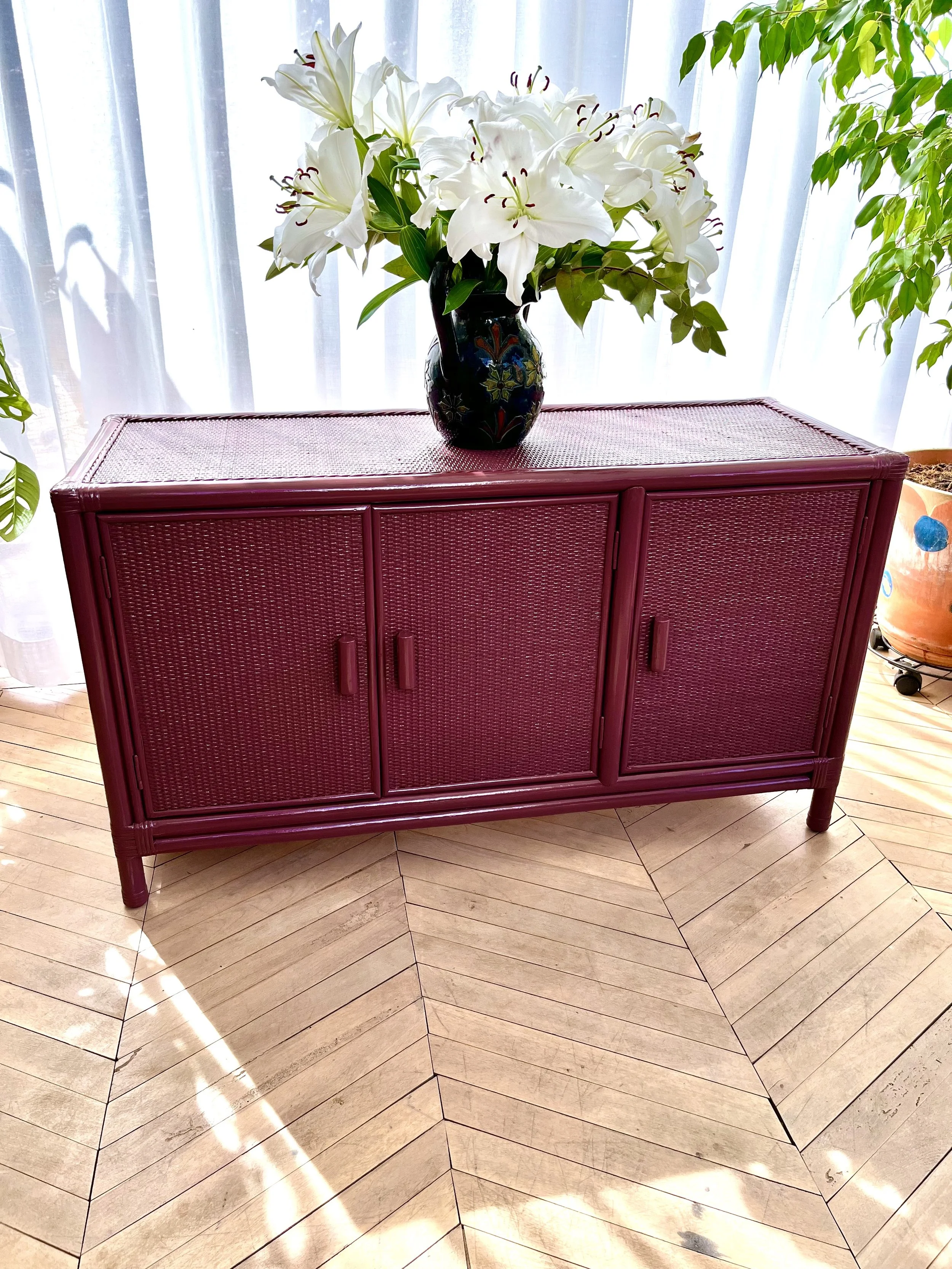 A pink cabinet with a textured surface is positioned on a parquet floor, with a black vase filled with white lilies and green leaves placed on top, set against a backdrop of white curtains and houseplants.