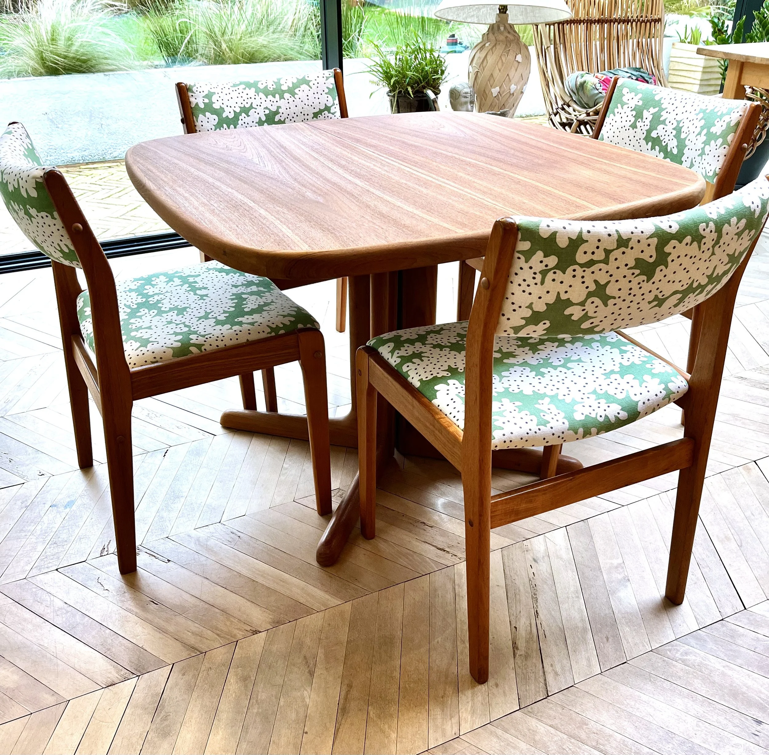 A wooden dining table with four matching wooden chairs with patterned cushions, situated near large glass windows with a view of green plants outside.