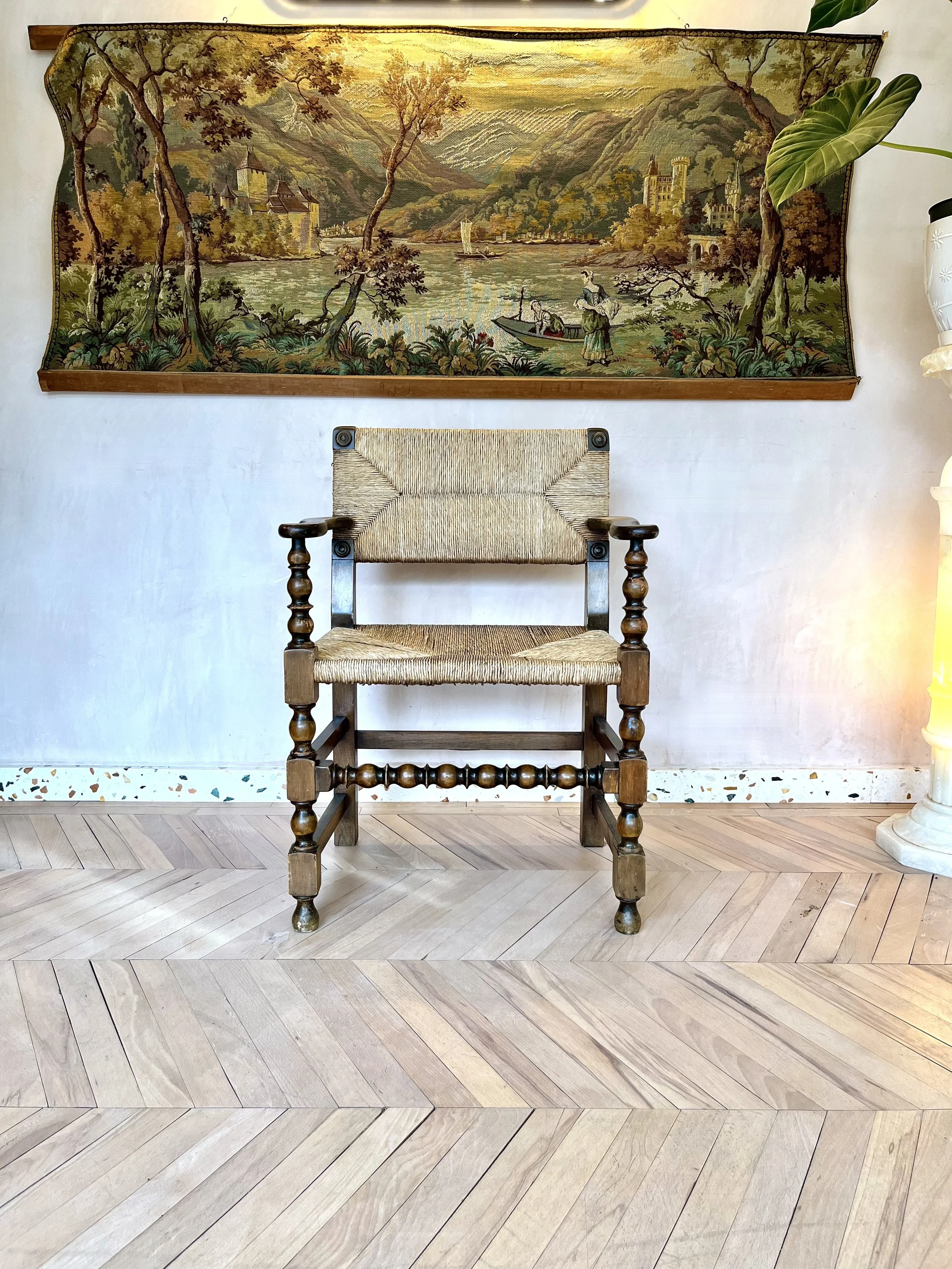 A woven wooden chair with armrests and turned legs centered in front of a landscape tapestry depicting trees, mountains, a river, and castles, hung on a white wall. To the right of the scene is a large green leafy plant in a white pot, and to the low