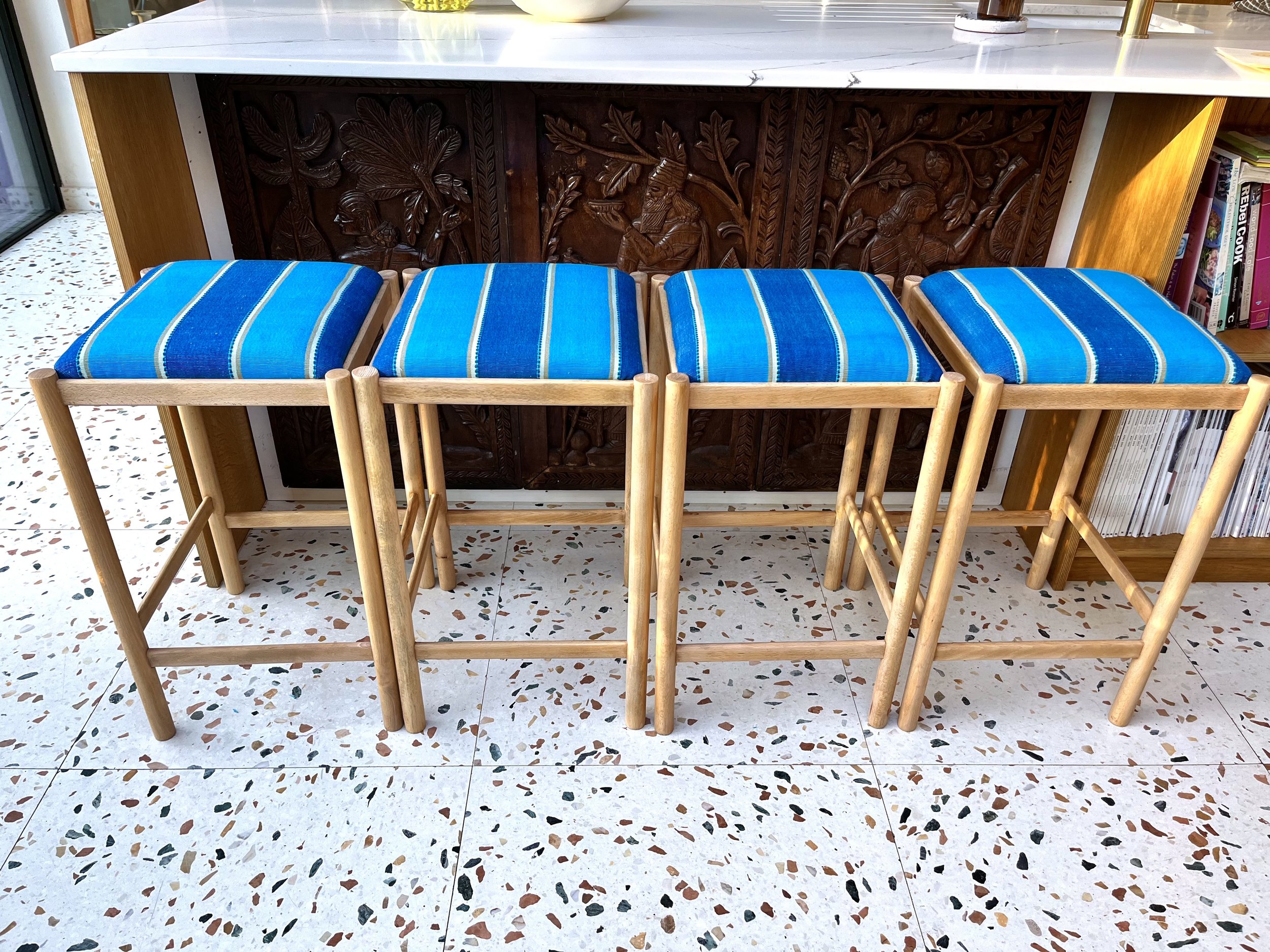 Four wooden chairs with blue striped cushions placed in front of a dark carved wooden panel, part of a piece of furniture, with a white marble countertop above. To the right, a bookshelf with several books is visible.