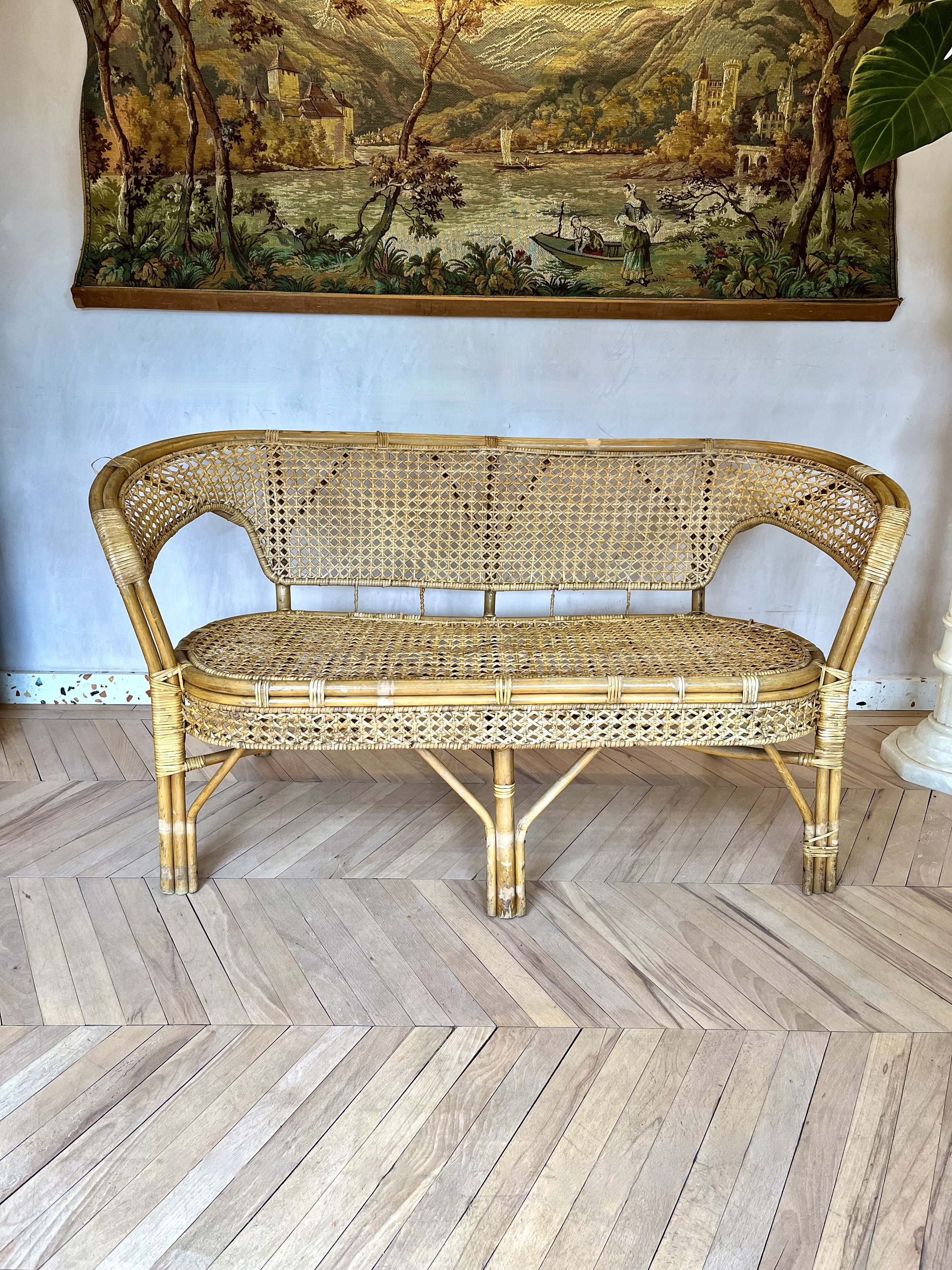 A woven rattan bench with a curved backrest and armrests, placed on a light wood-patterned floor. Behind it is a large, framed tapestry depicting a scenic landscape with a river, trees, a boat with two women, and castles in the background.