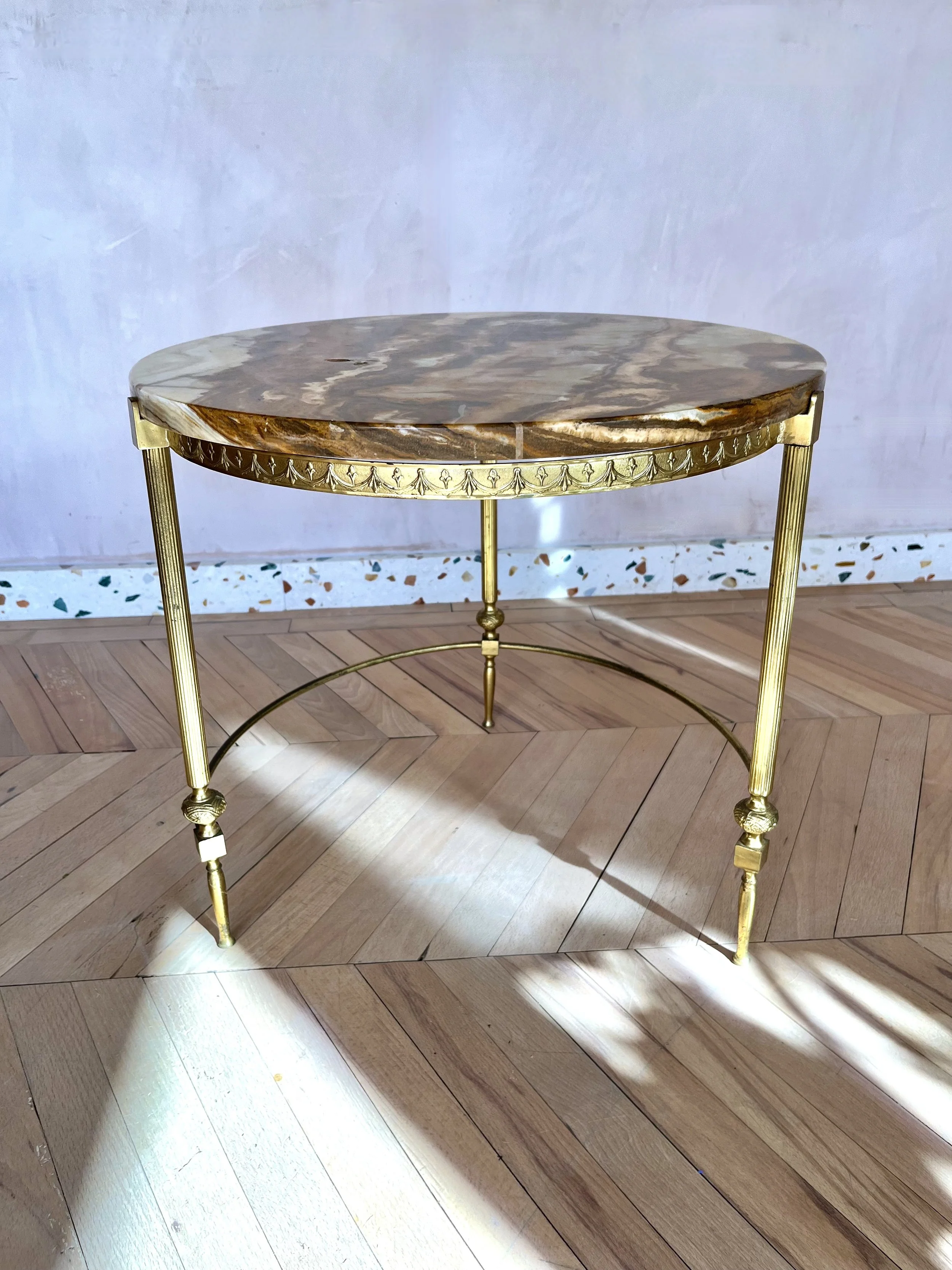A round table with a marble top and gold legs standing on a wooden floor against a light-colored wall.