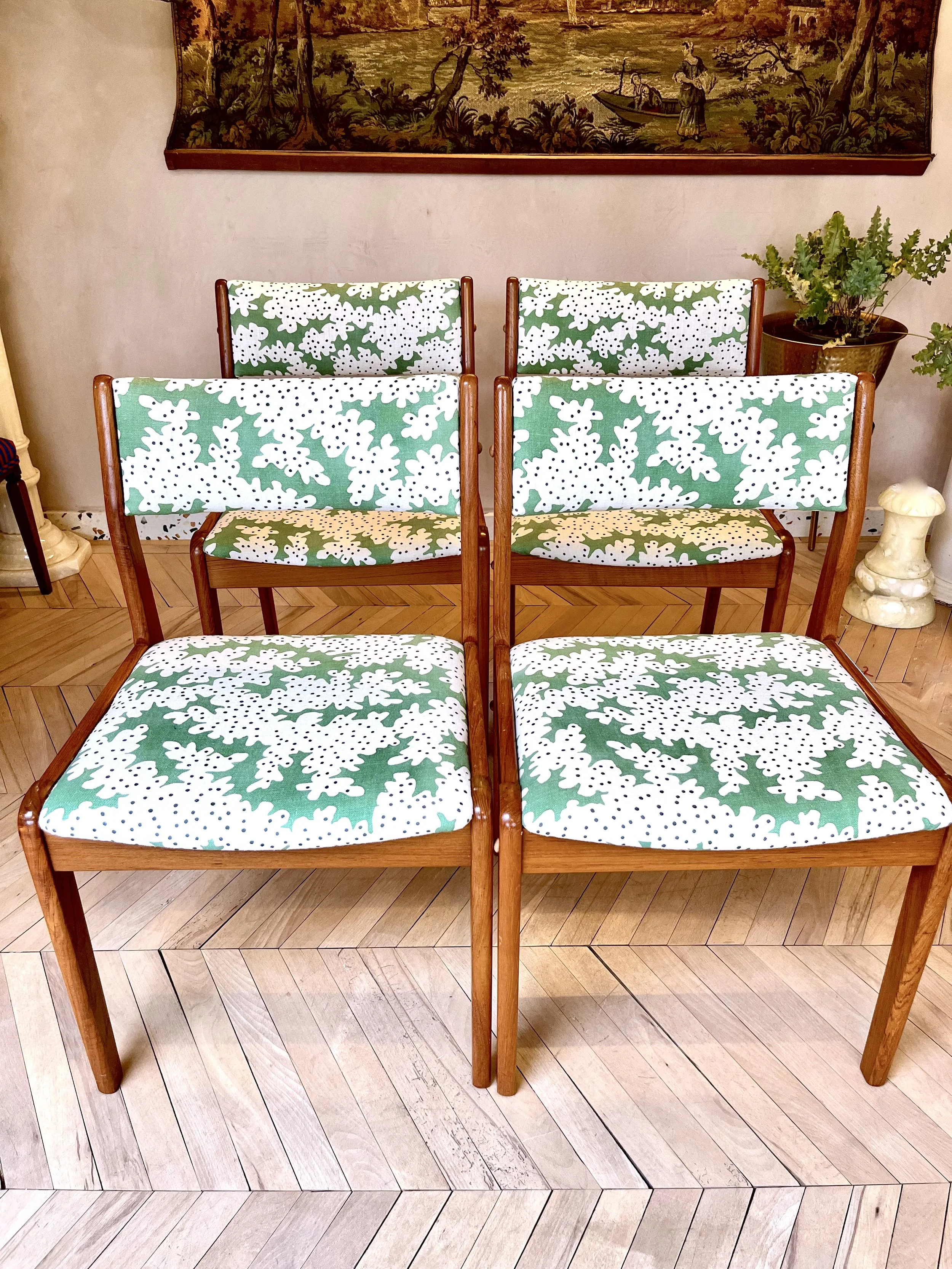Four wooden chairs with cushions featuring green holly and white snowflake patterns in a room with a beige wall, a tapestry depicting a nature scene with trees and people in a boat, a potted plant on the right, and a wooden floor.