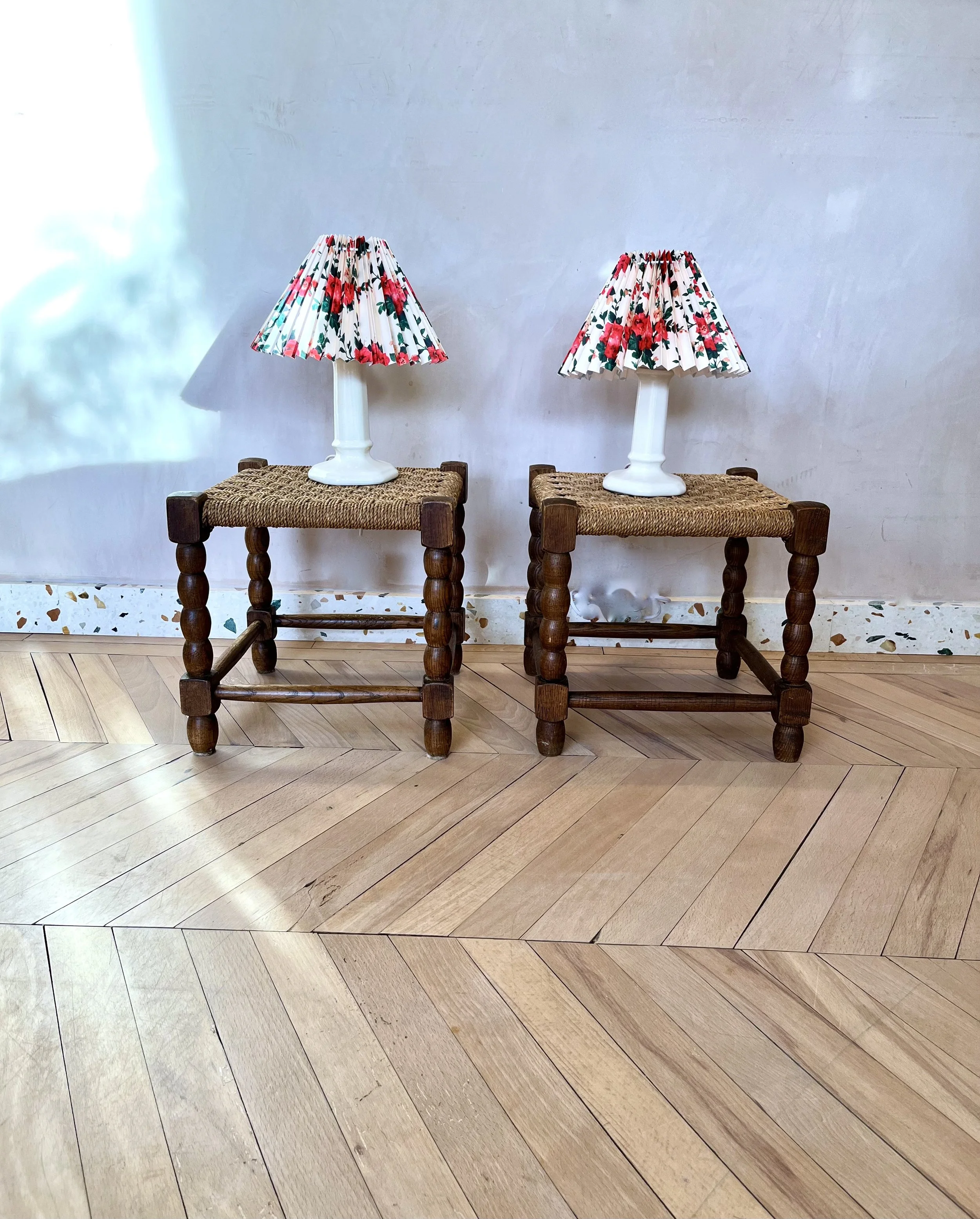 Two bedside lamps with floral patterned shades are placed on woven surface stools in front of a plain white wall, with a patterned wooden floor below.