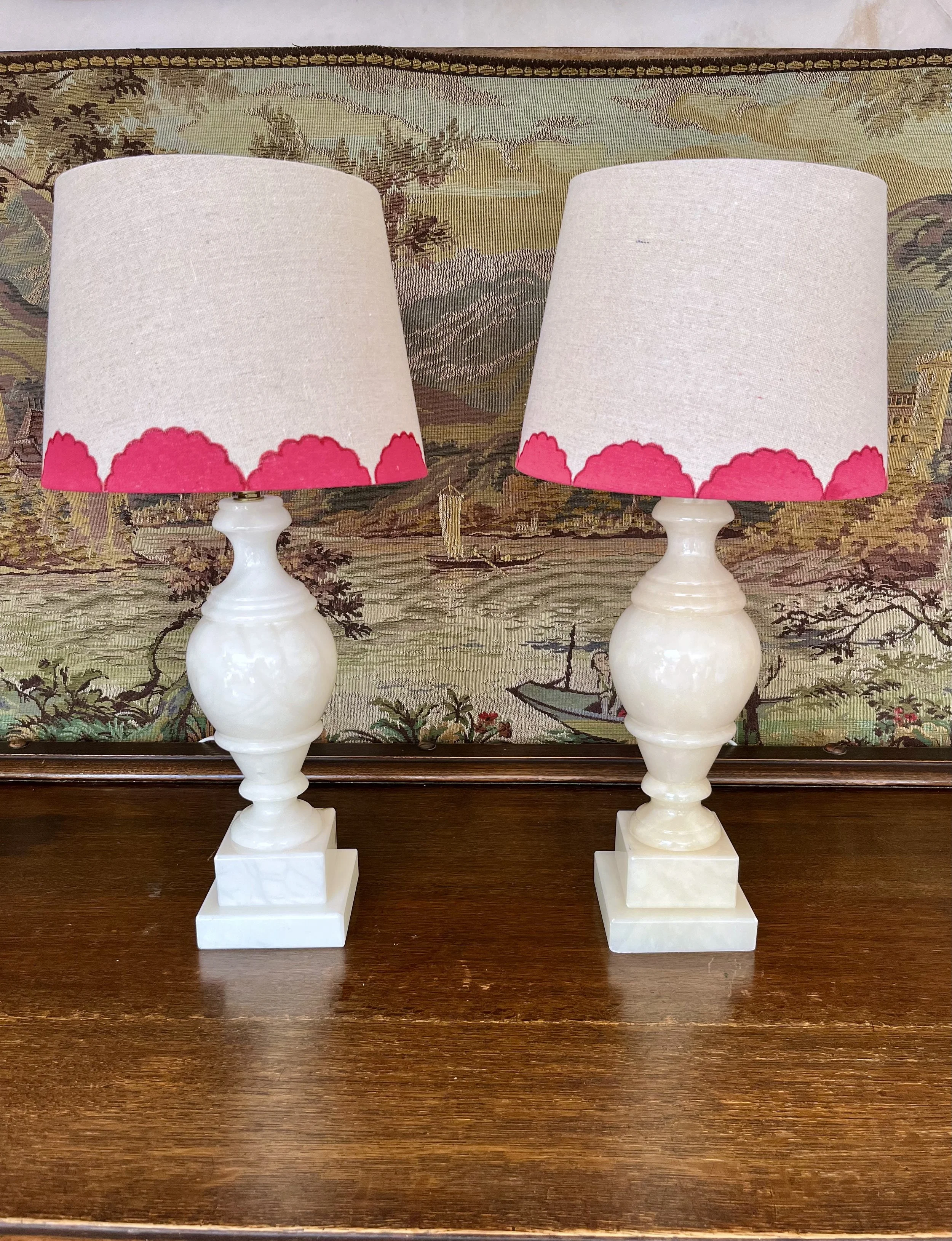Two white ceramic table lamps with pink petal-patterned lampshades, placed on a polished wooden surface with a decorative tapestry in the background.