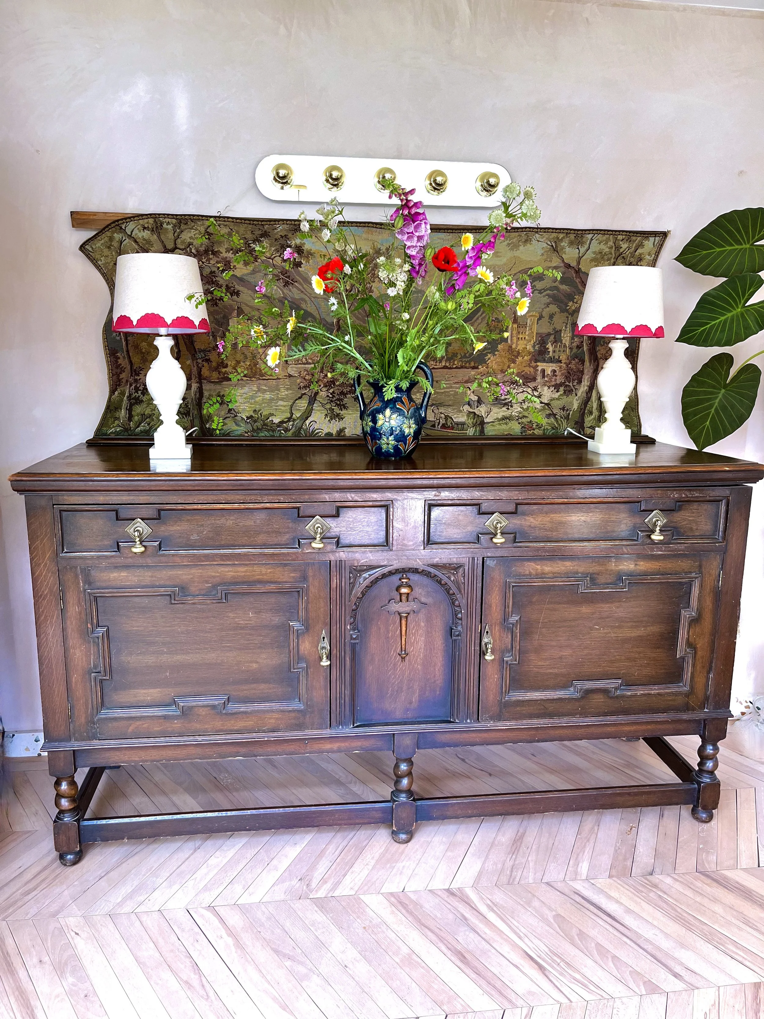 A vintage wooden sideboard with a dark finish, ornate details, and rounded legs. On top, there are two white lamps with pink scalloped trim and a large floral arrangement in a dark blue vase with floral patterns. Behind the arrangement, there is a ta