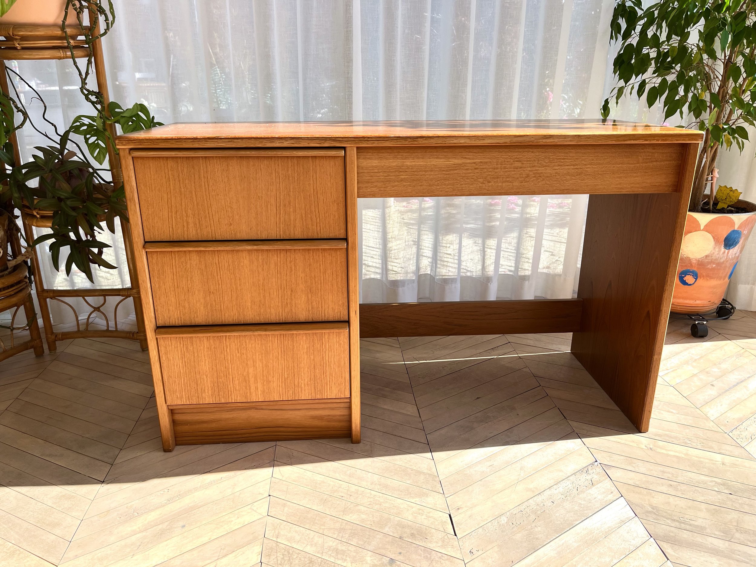 A wooden desk with a natural finish positioned in front of sheer white curtains. To the left, there is a plant in a woven basket stand, and to the right, a colorful decorative pot with a plant. The desk has a sliding drawer on the right side.