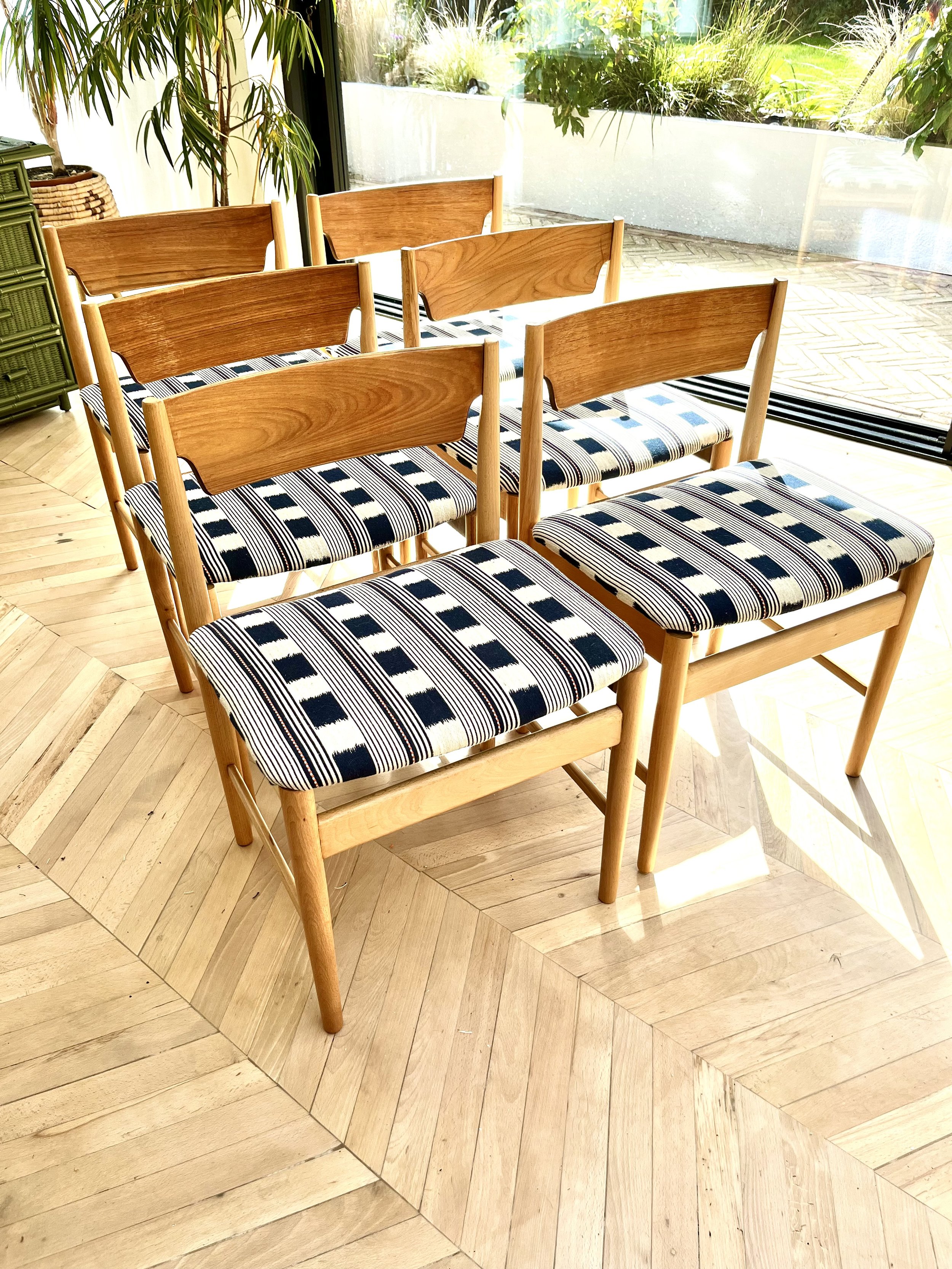 Wooden chairs with striped fabric cushions arranged in a room with large glass windows and hardwood flooring, overlooking a garden with plants outside.