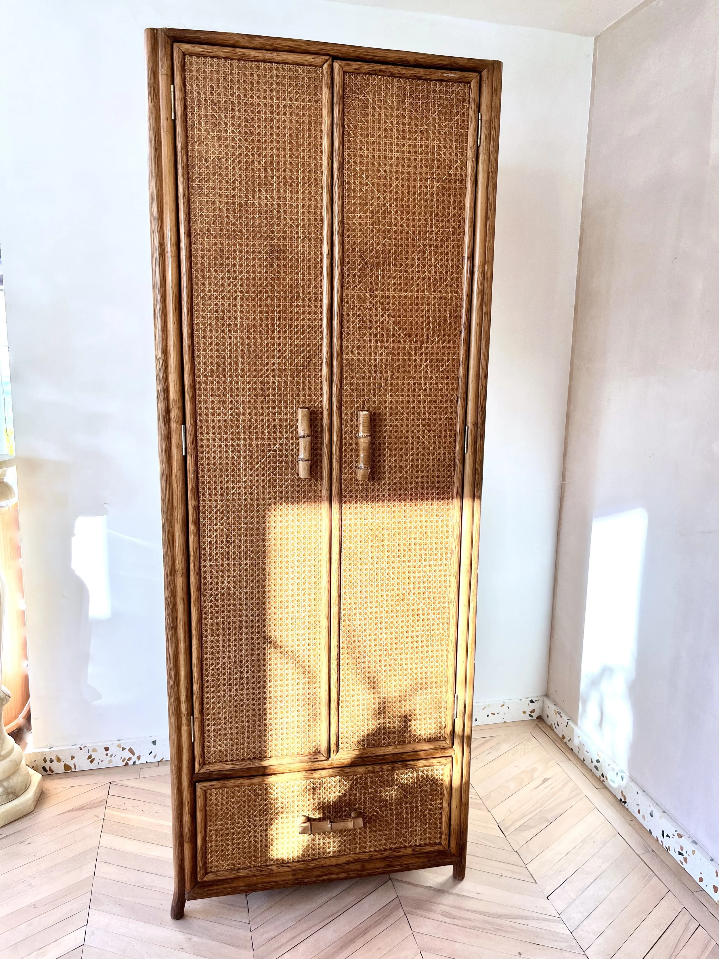 A wooden cabinet with woven cane doors and handles, standing in a room with light-colored walls and parquet flooring.