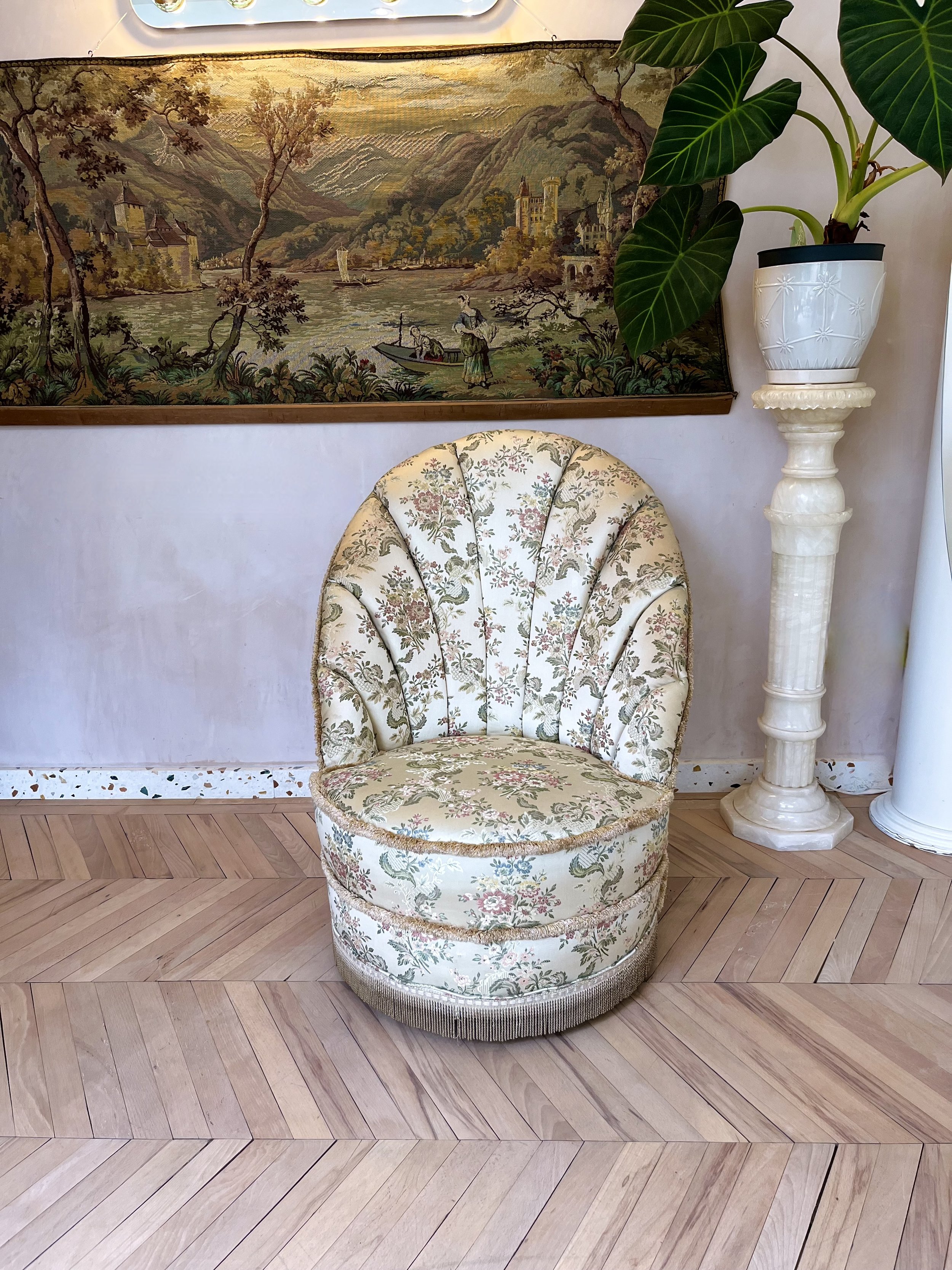 A vintage upholstered armchair with floral fabric and fringe trim at the bottom, placed on a hardwood floor with a herringbone pattern. Behind the chair, there is a large framed tapestry depicting a serene landscape with a river, trees, mountains, ca