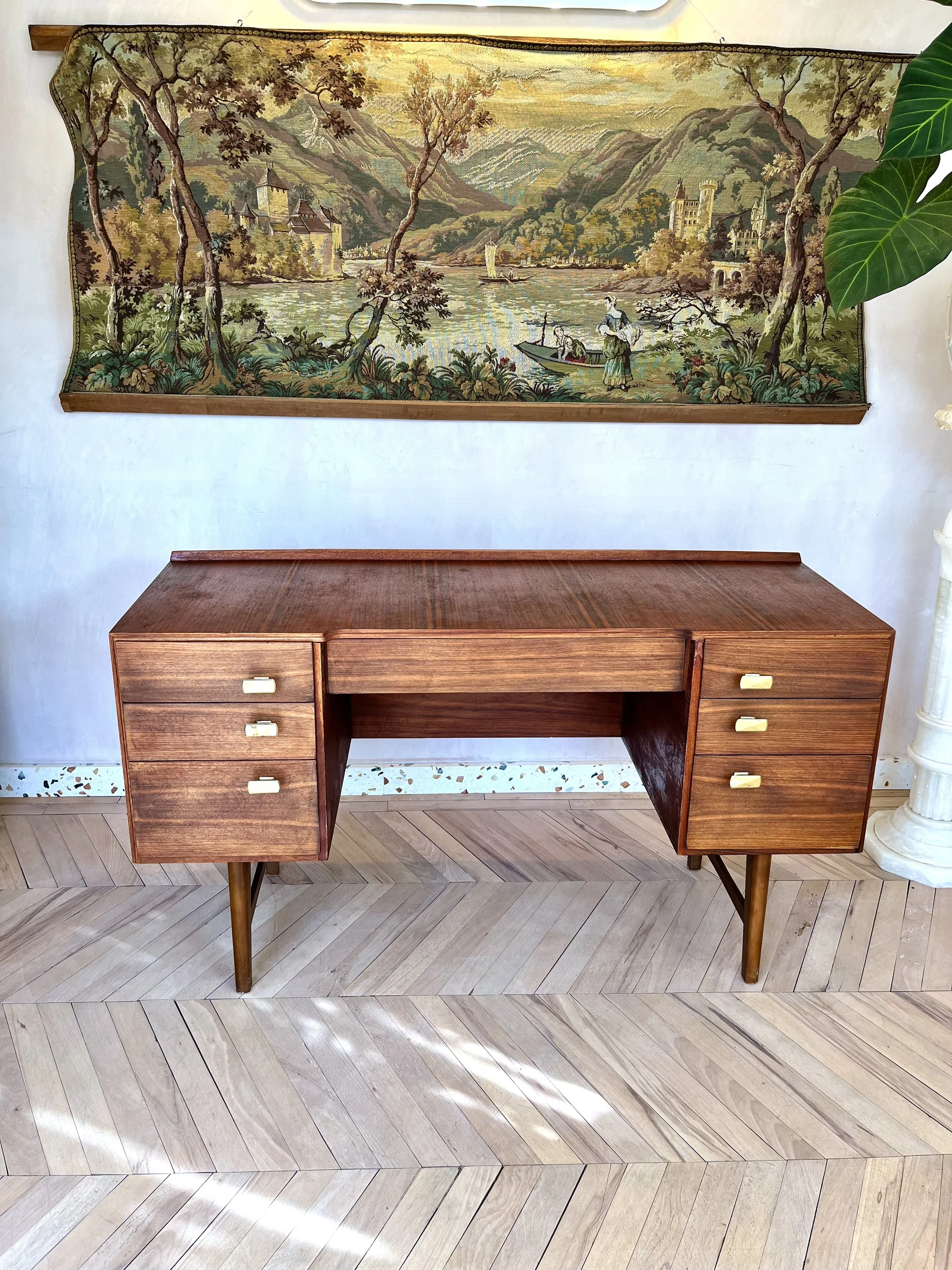 Wooden desk with a flat surface and six drawers, three on each side, standing on angled legs in a room with a wooden patterned floor. A large tapestry with a landscape scene hangs on the wall behind it.