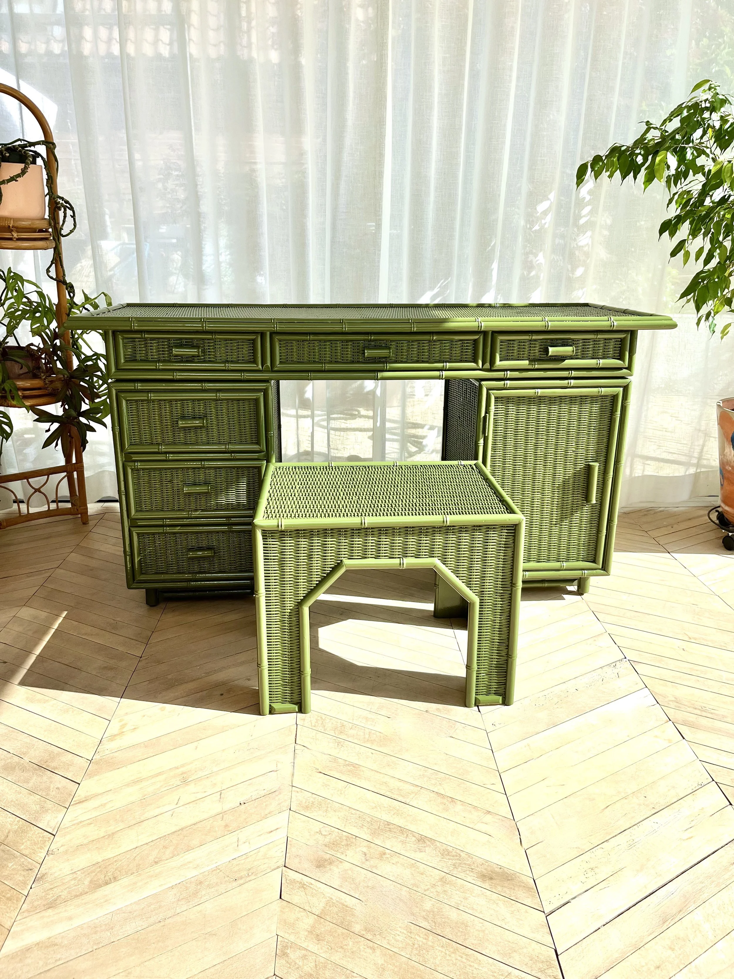 Green wicker furniture piece set against a sheer white curtain with plants on either side, on a light wood floor.