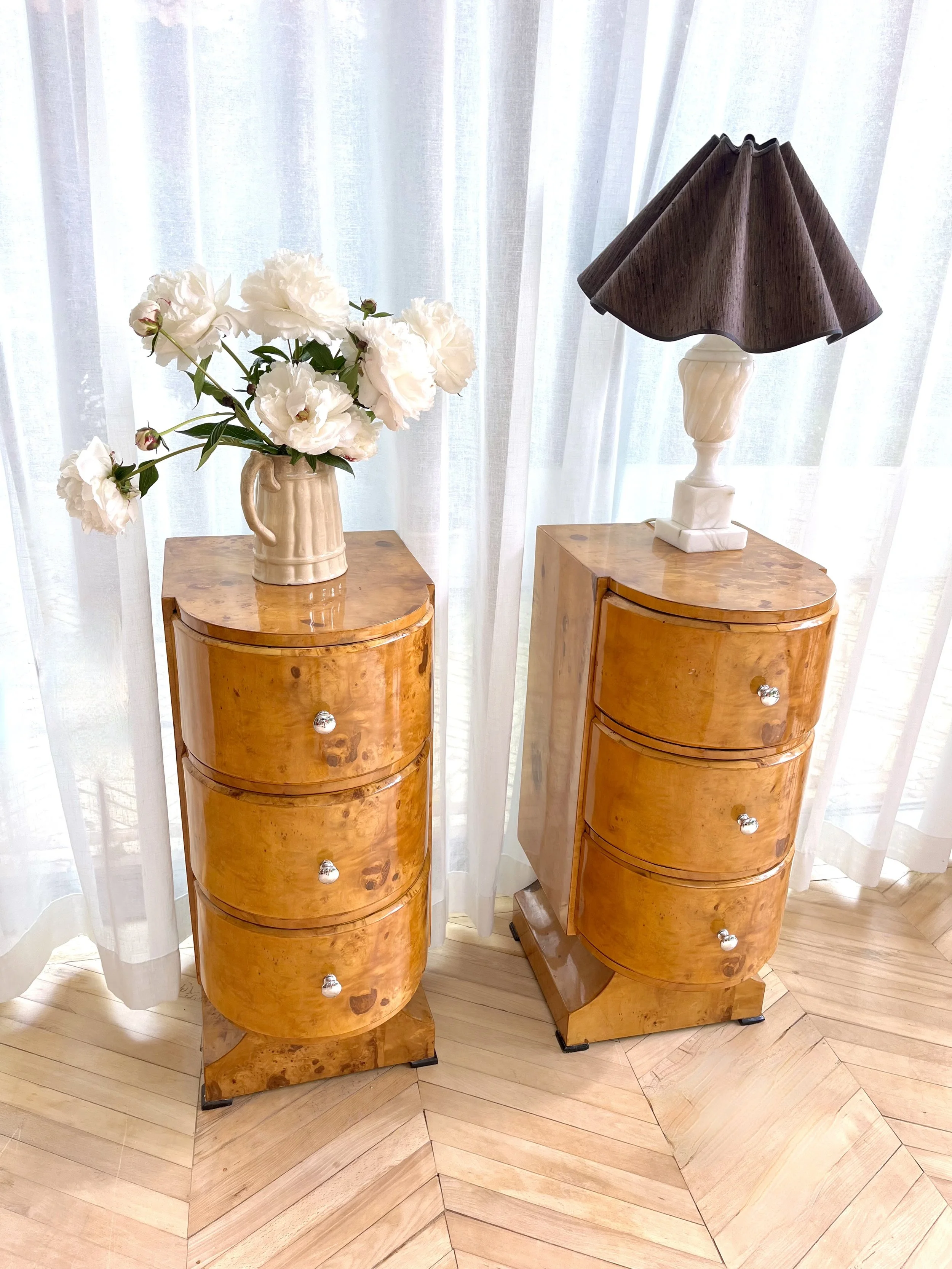 A pair of vintage wooden bedside tables with a glossy finish and metallic knobs, placed on a herringbone wood floor, with a set of white curtains in the background. One table has a ceramic vase with white peony flowers, and the other has a cream-colo