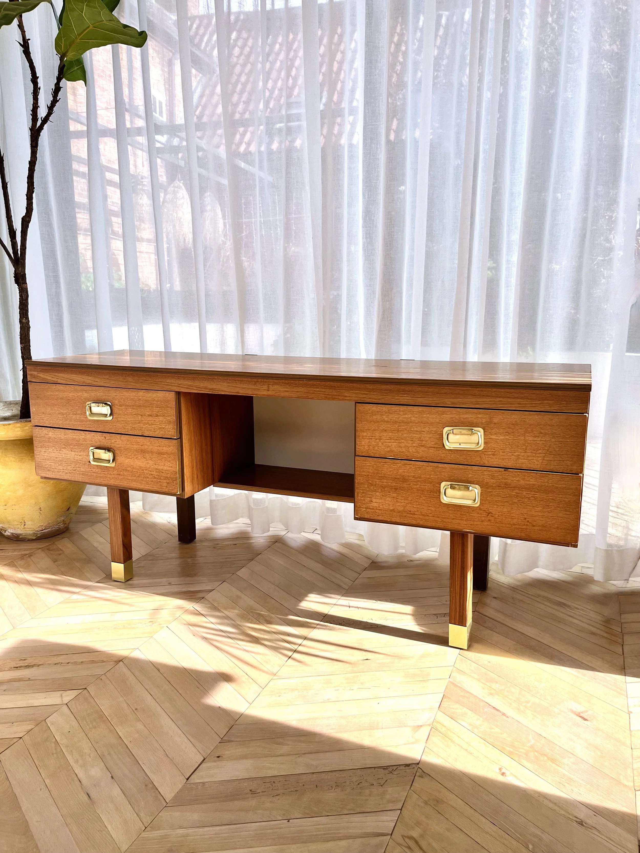 A wooden desk with four drawers and a central open space, placed on a wooden herringbone floor, with white sheer curtains behind it letting in natural light. To the left, there is a tall potted plant with big green leaves.