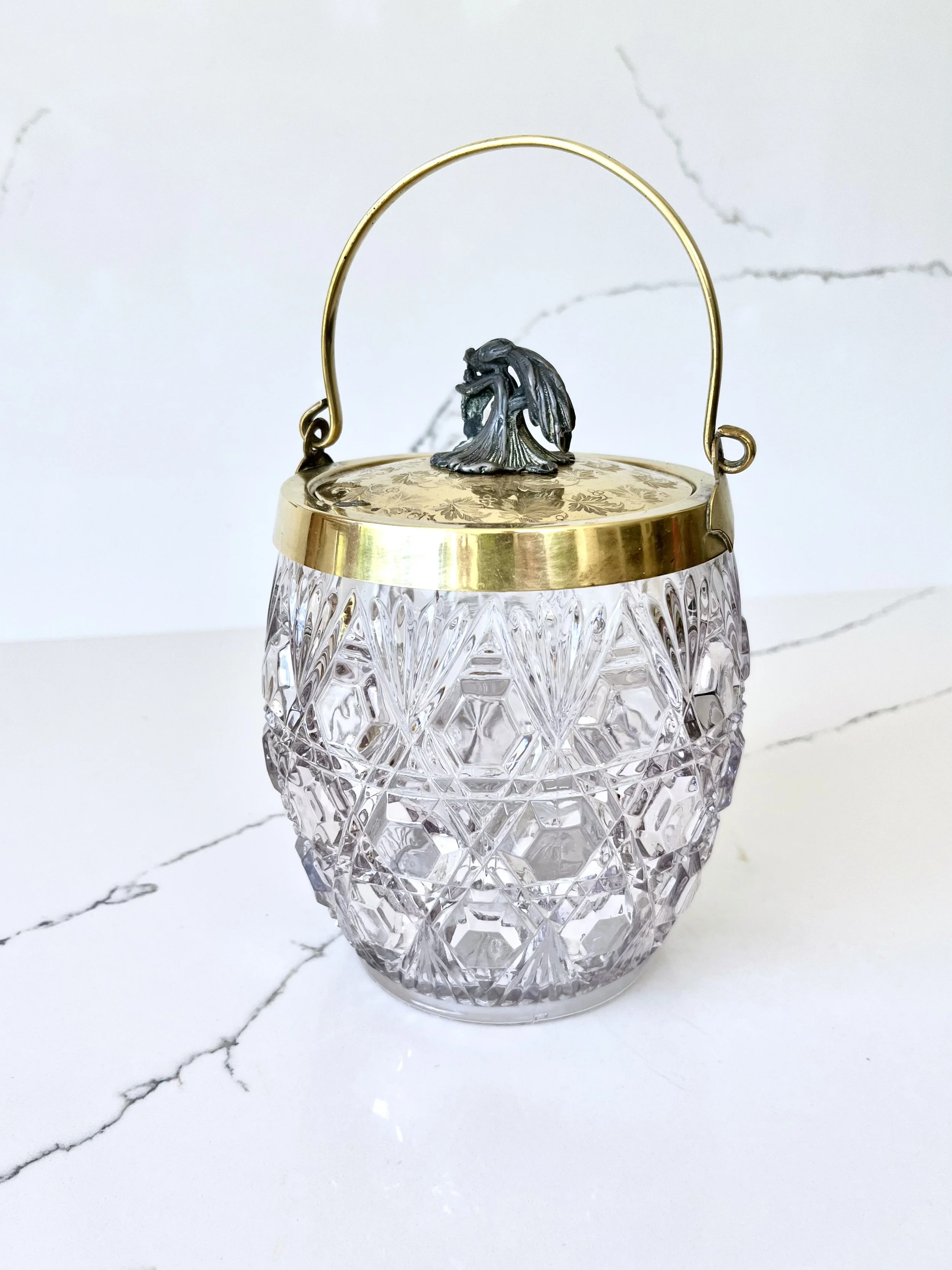 A crystal glass container with a patterned surface, topped with a gold lid and a decorative metal handle featuring a small animal figure.