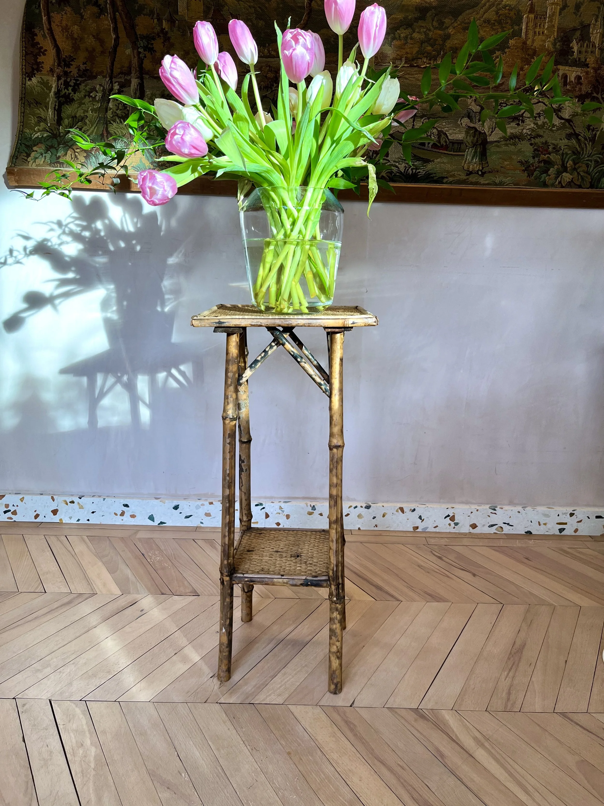 A bouquet of pink and white tulips in a clear glass vase on a wooden stand, with a tapestry hanging on the wall behind.