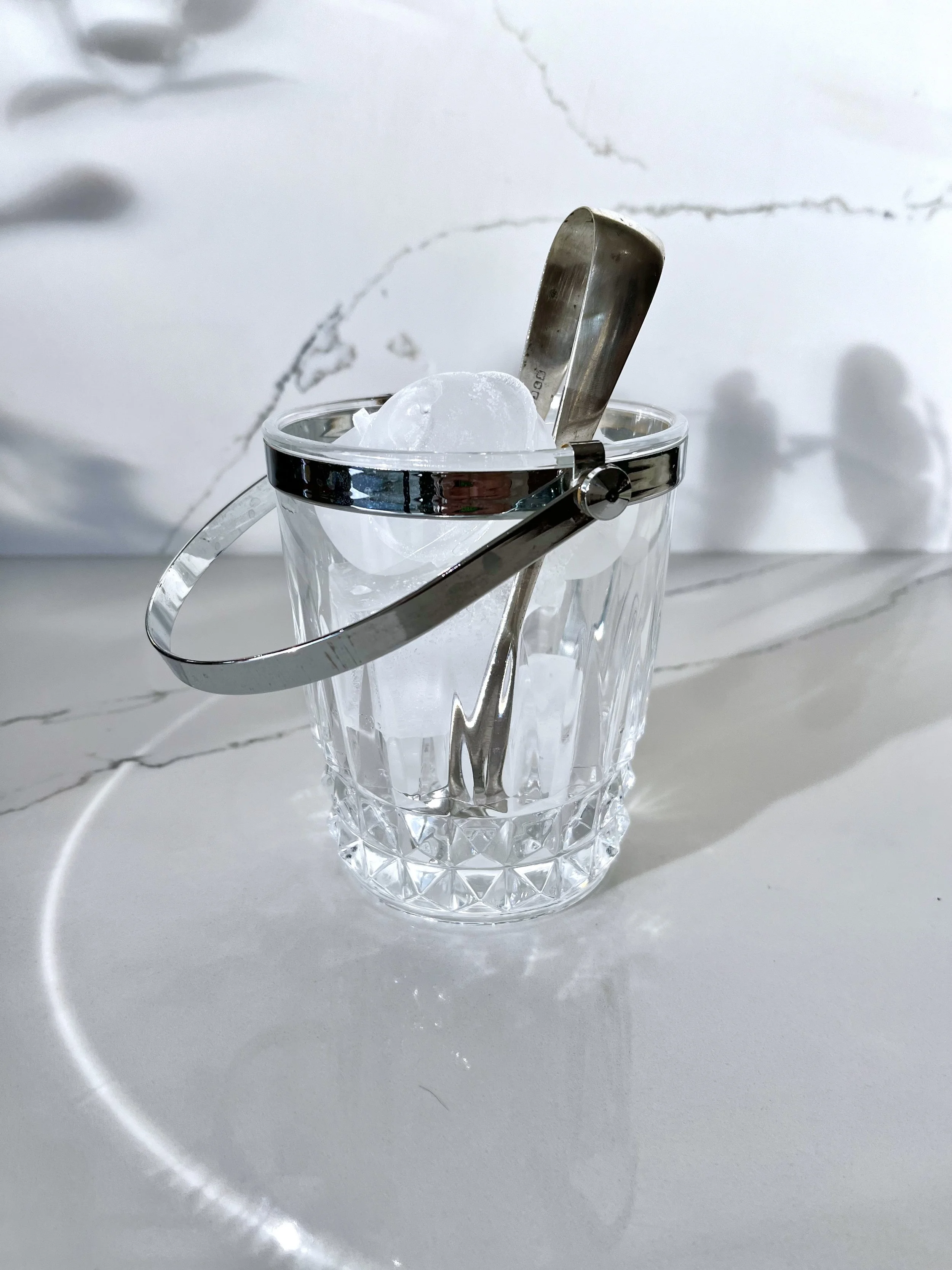 A clear glass tumbler with geometric patterns on its surface, filled with ice cubes. A pair of metal ice tongs and a metal ice scoop are resting on the top of the ice, with a white marble surface in the background.