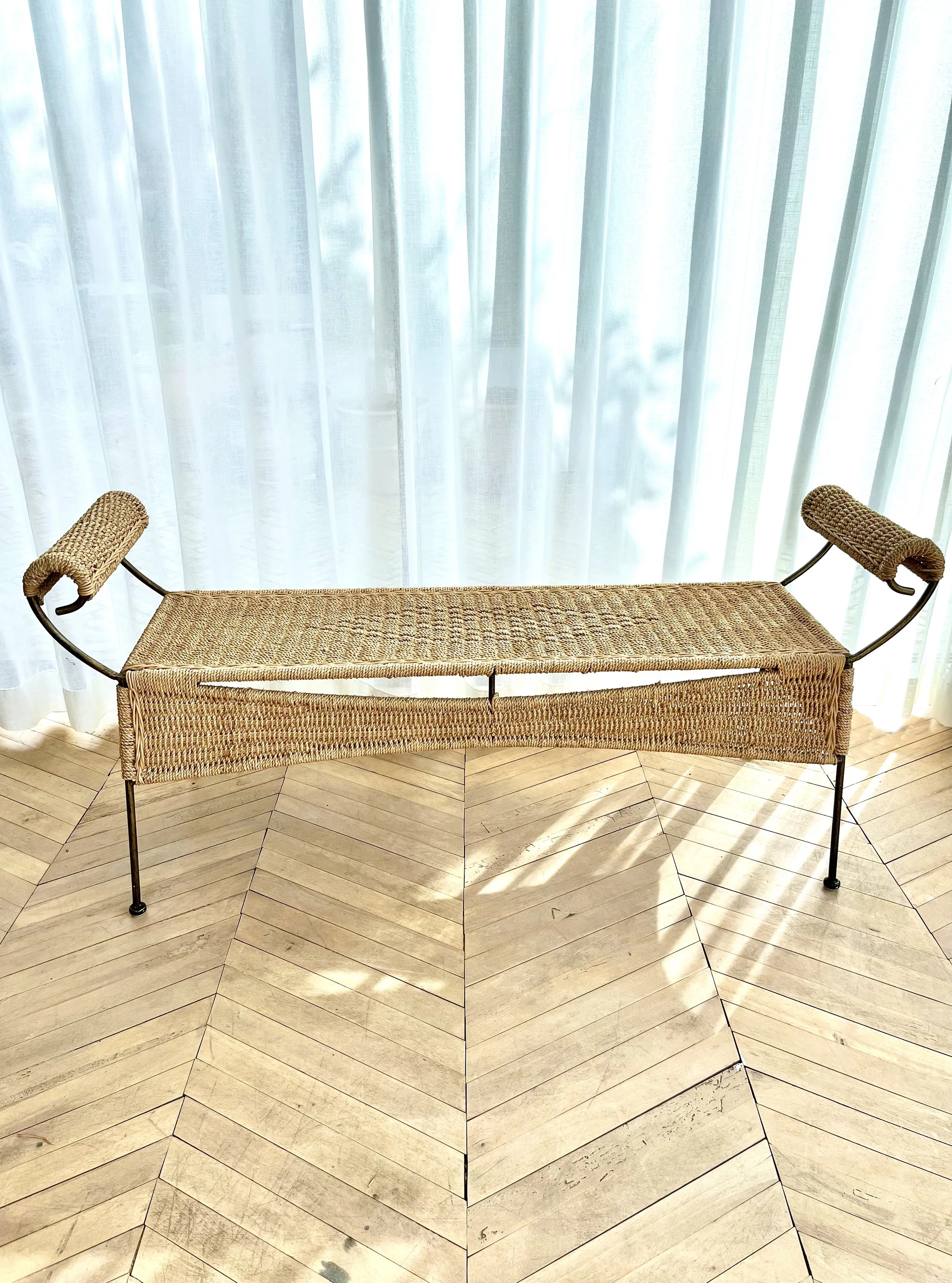 A woven wicker bench with a metal frame and curved armrests is placed on a wooden chevron-patterned floor in front of sheer white curtains.