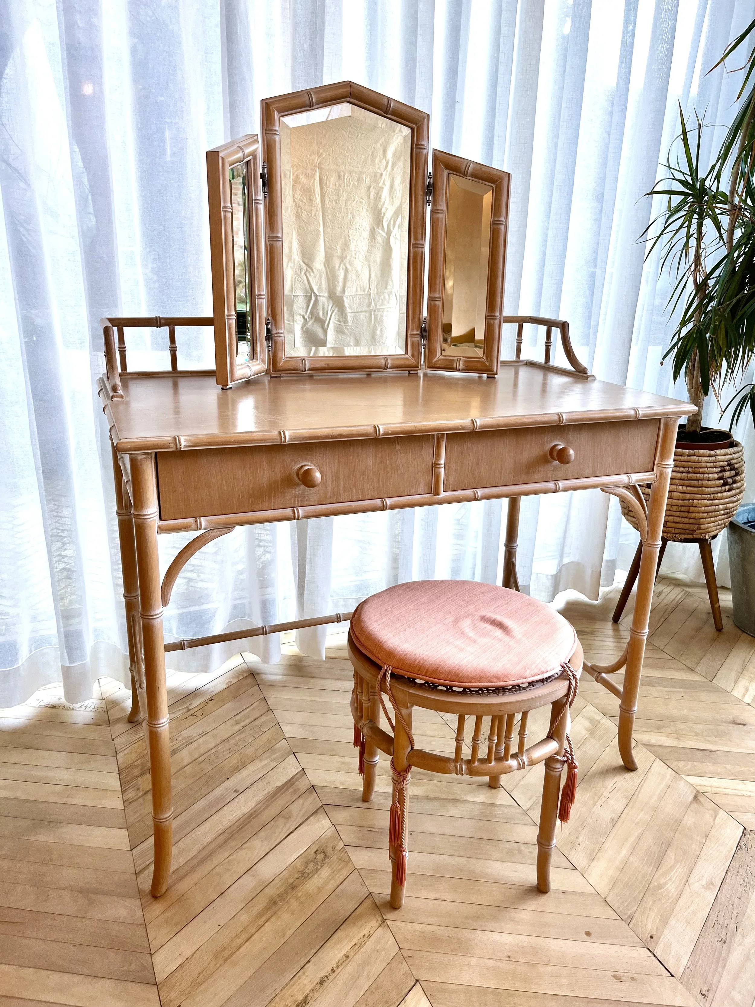 A wooden dressing table with three mirrors in a room with sheer curtains and wooden flooring. An orange cushioned stool is in front of the table, and a tall potted plant is on the right.