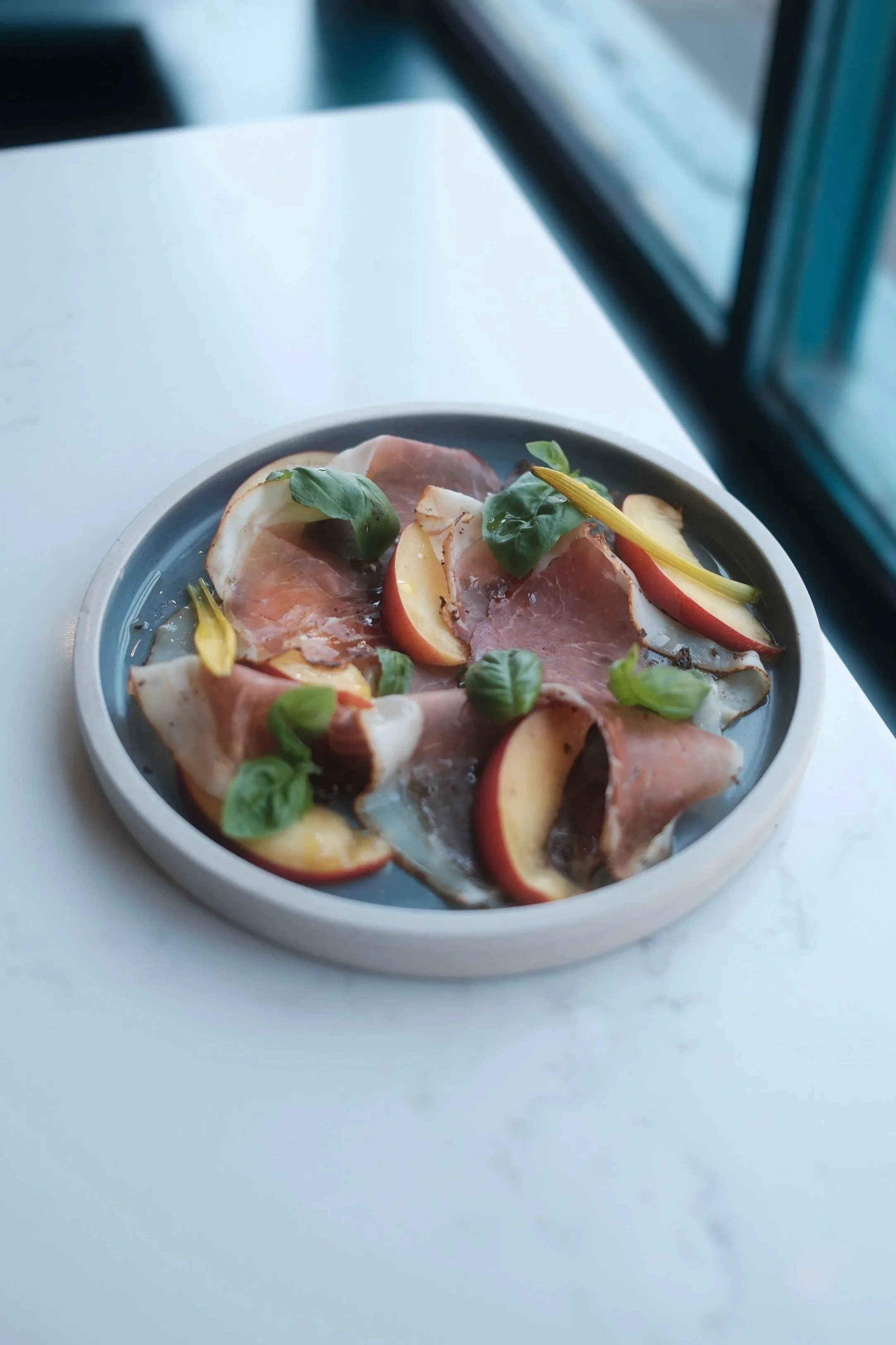 A plate of thinly sliced prosciutto, apple slices, fresh basil leaves, and yellow flower petals on a gray dish, placed on a white surface near a window.