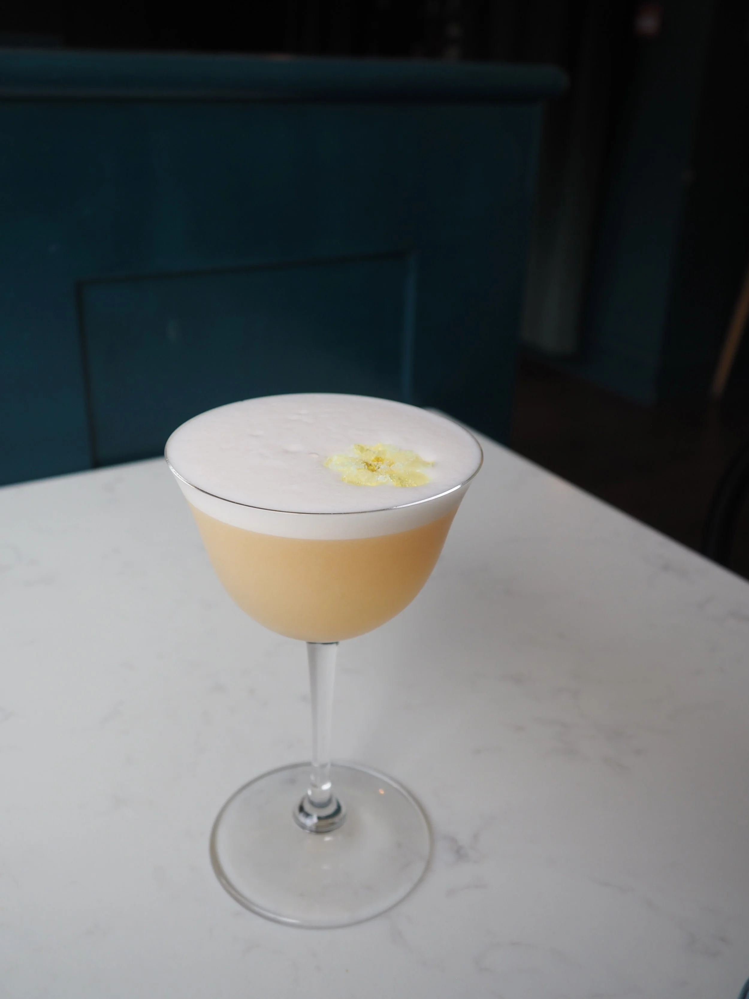 A cocktail in a tall, elegant glass with a white frothy top garnished with a lemon peel peel on top of the foam.